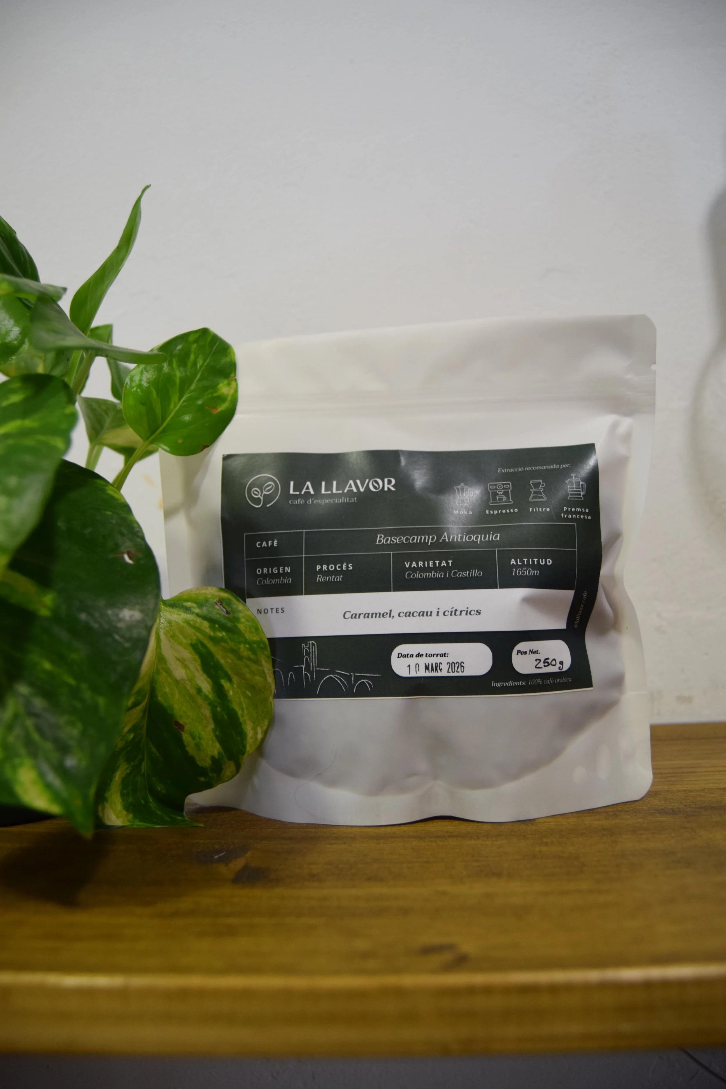 Paquete de café La Llavor en una mesa de madera, próximo a una planta con hojas verdes y amarillas.