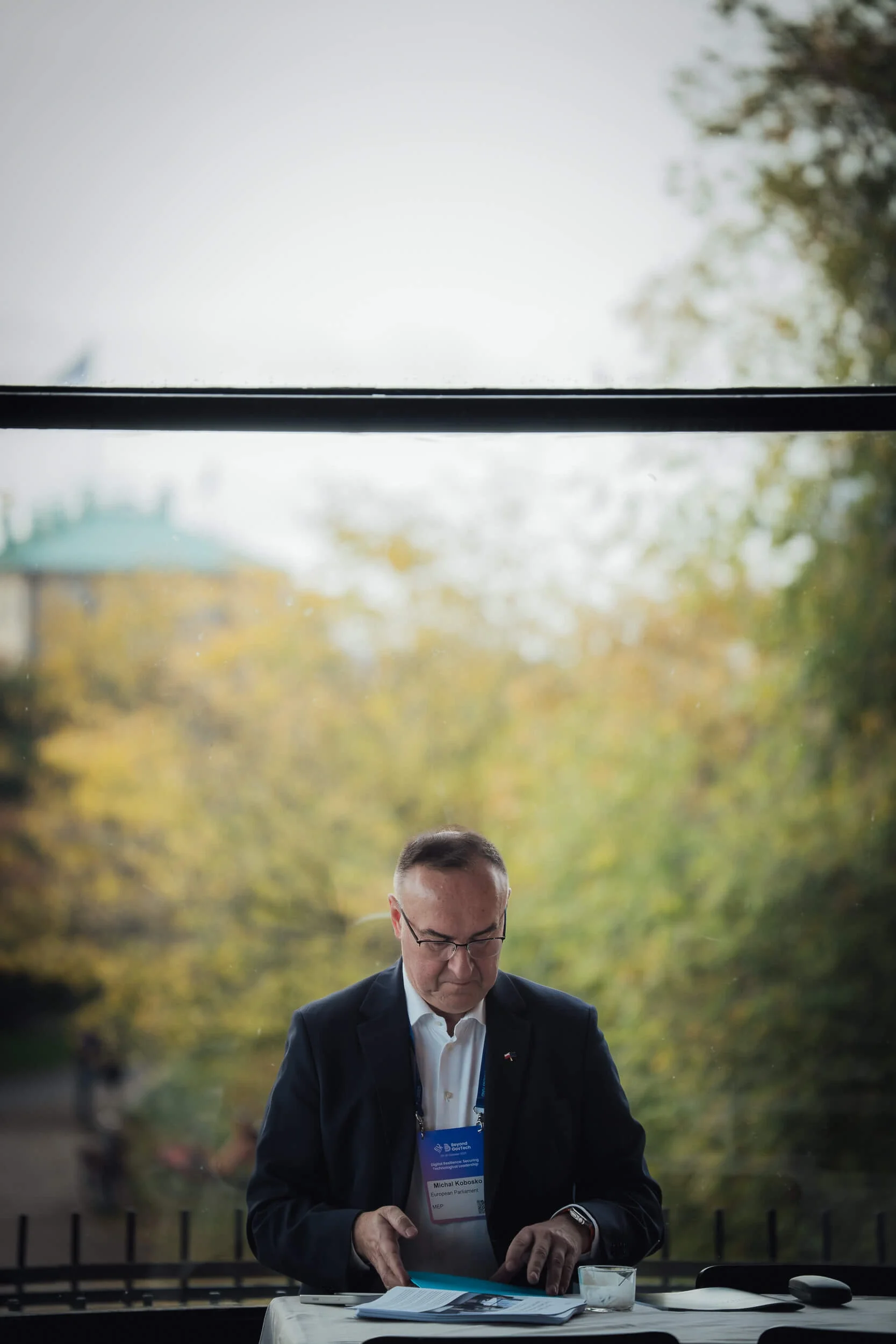 En mand til Beyond GovTech i jakkesæt med navneskilt sidder ved et bord indendørs, læsende dokumenter foran ham, med en grøn park i baggrunden og træer med efterårsfarvede blade.