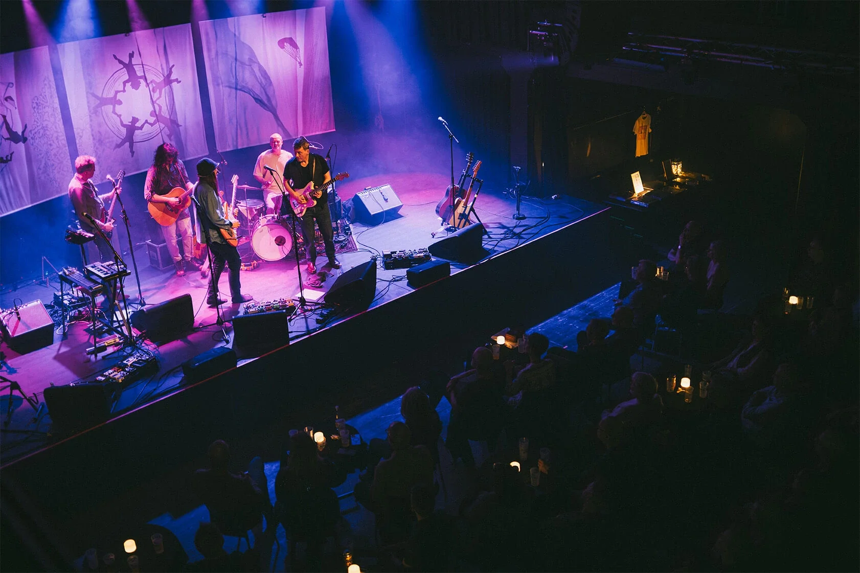 En musikgruppe optræder live på scenen hos Fermaten med blå og lilla belysning, mens publikum ser på. Baggrundsbilledet har dansende silhuetter og et flag. Publikum sidder på stole med lyskilder på borde.