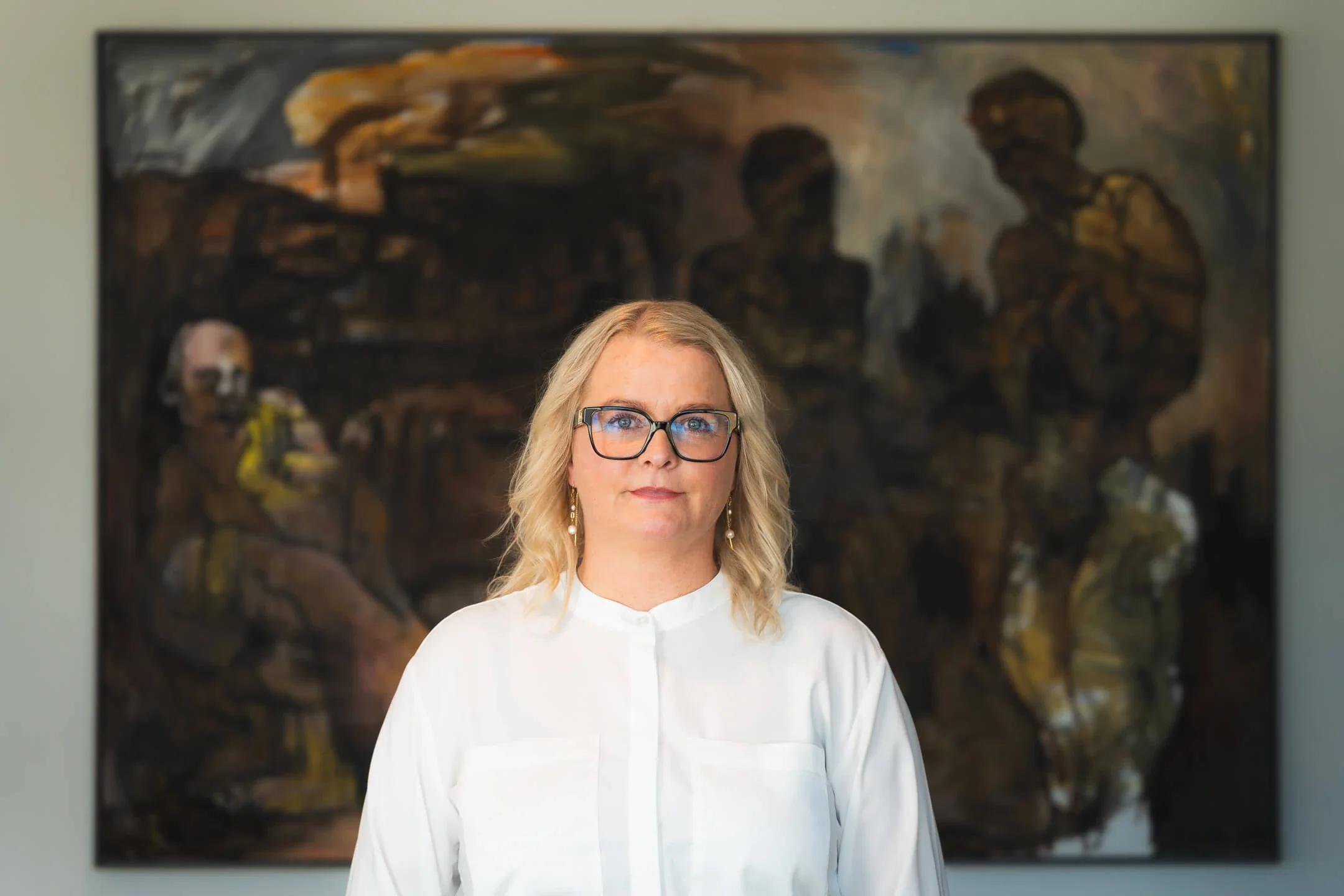 Portræt af en kvindelig formand fra HK MidtVest med blondt, langt hår og briller, iført en hvid skjorte, stående foran et kunstværk.
