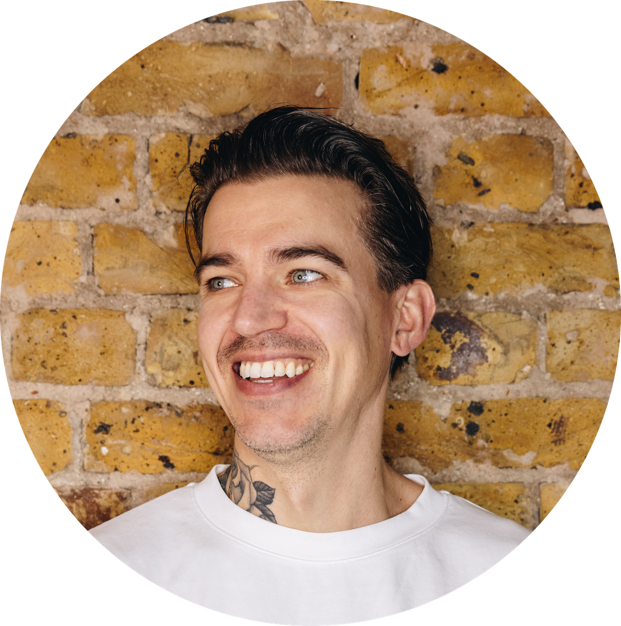 Portrait of a young man with dark styled hair, blue eyes, and a tattoo on his neck, smiling and looking to the side, with a brick wall in the background.
