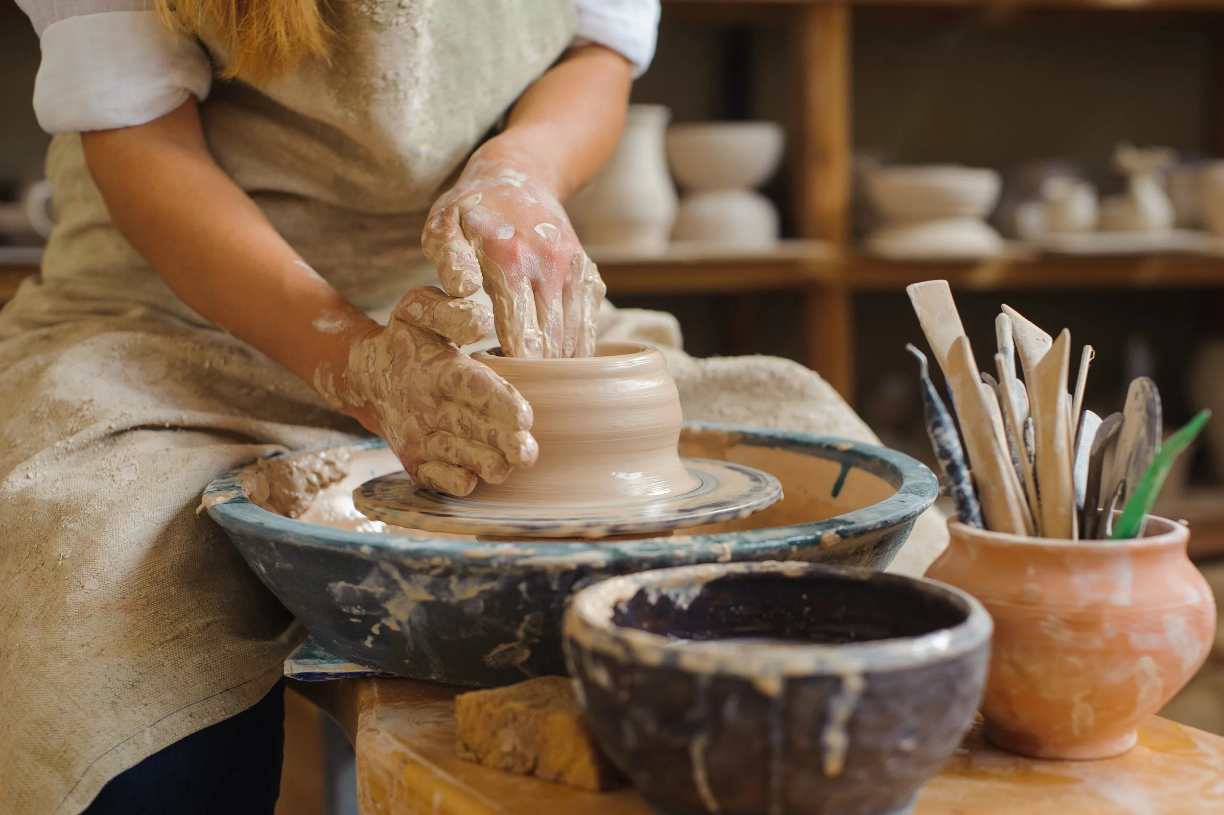 A person shaping a ceramic bowl on a pottery wheel in a pottery studio, surrounded by clay and pottery tools.