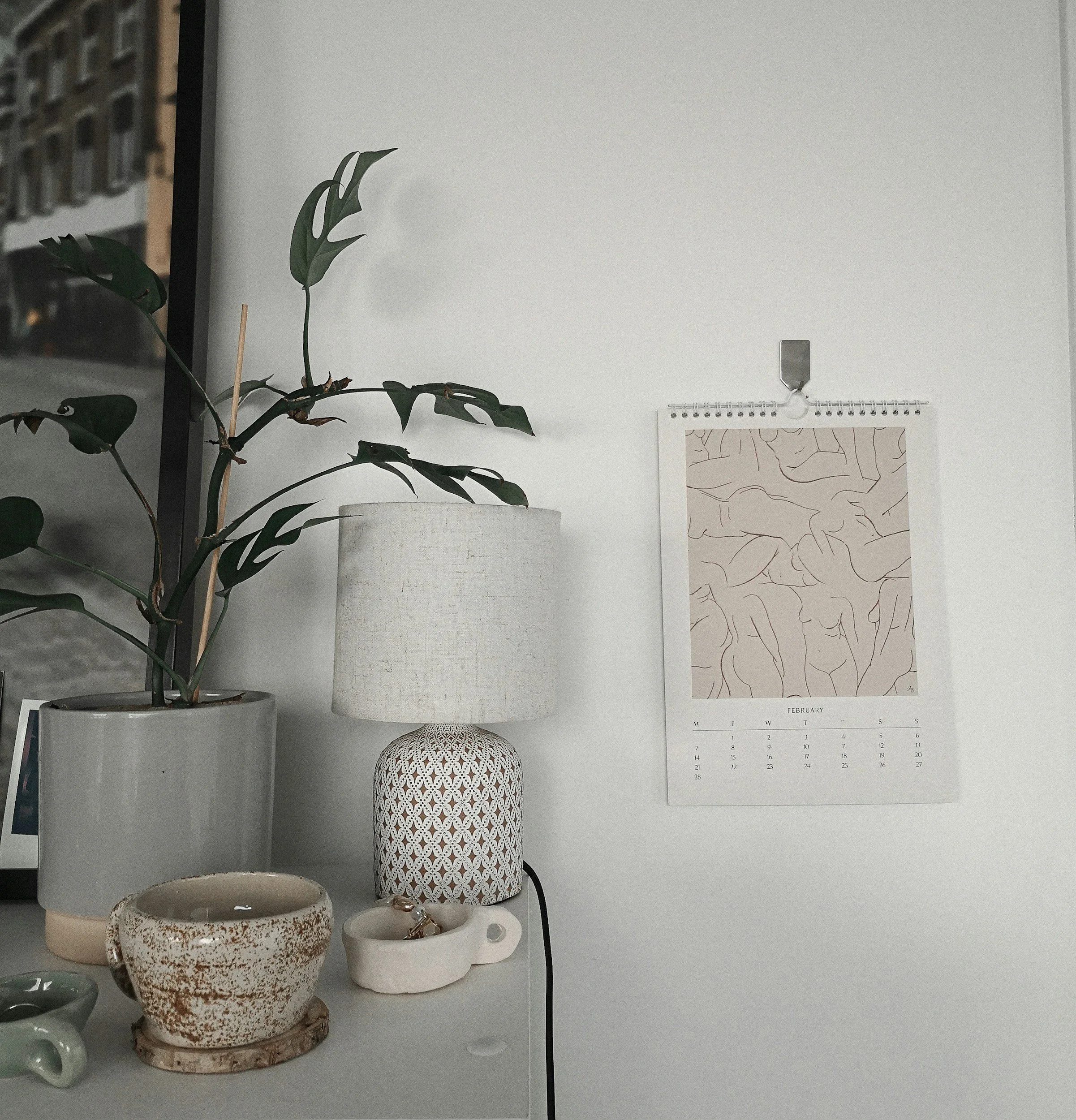 A corner of a room with a potted plant, a table lamp, and various ceramic decorative items on a surface. A wall calendar with a line art abstract image is hanging on a plain white wall.