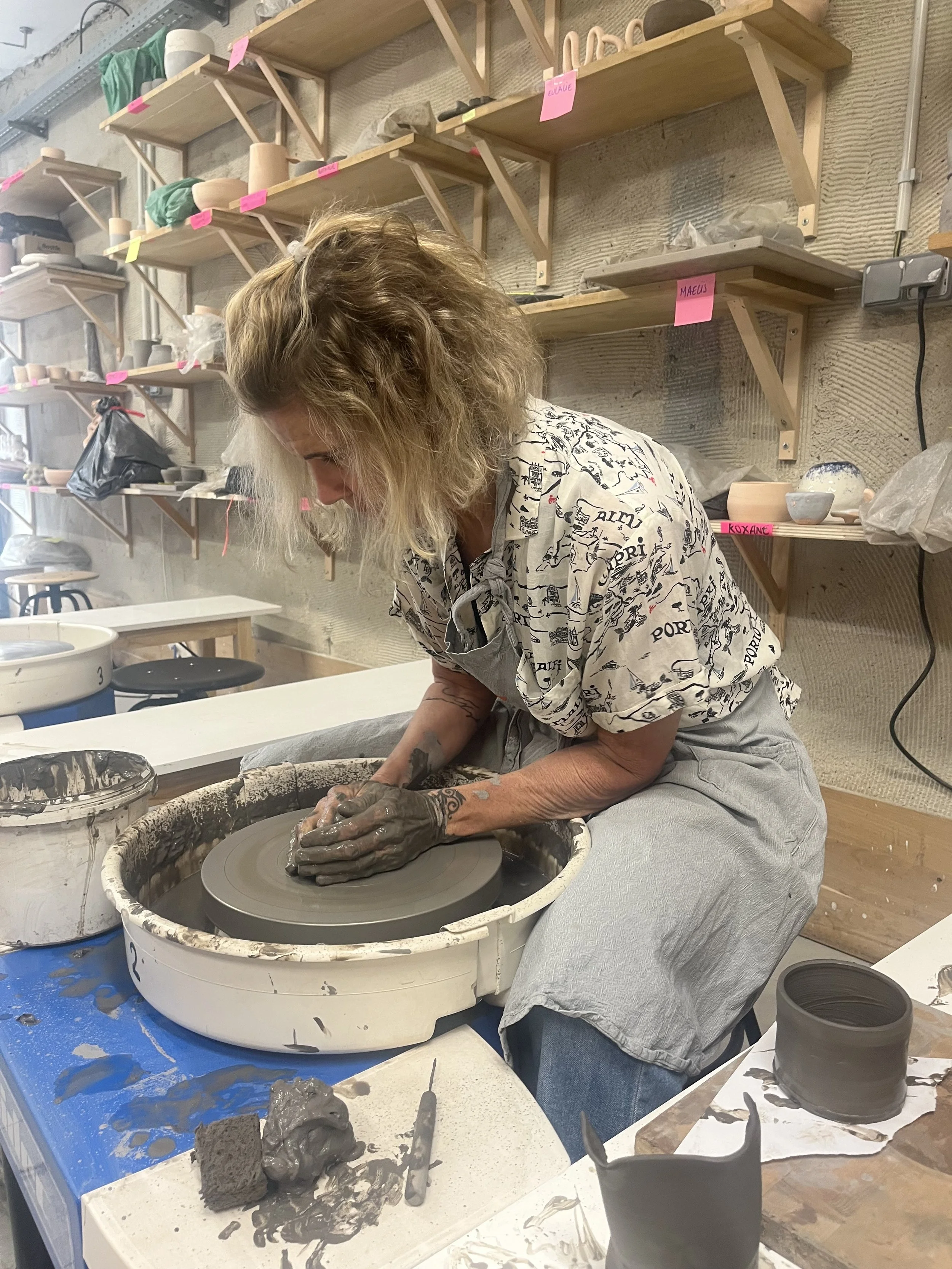 Woman shaping clay on a pottery wheel in a ceramics studio with shelves of supplies in the background.
