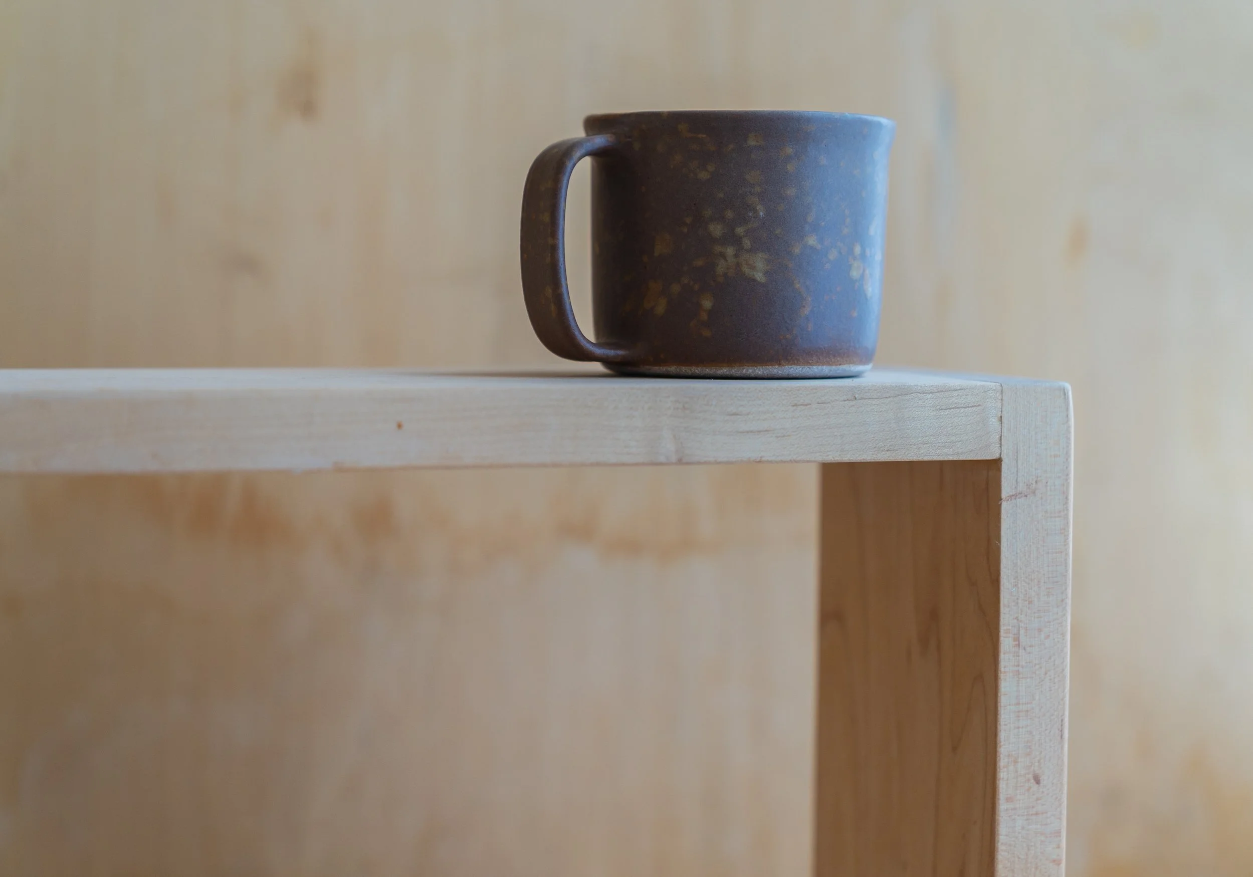 A rustic, dark brown ceramic mug with a speckled pattern sits on the corner of a light wooden table. The background features a plain, light-colored wall.
