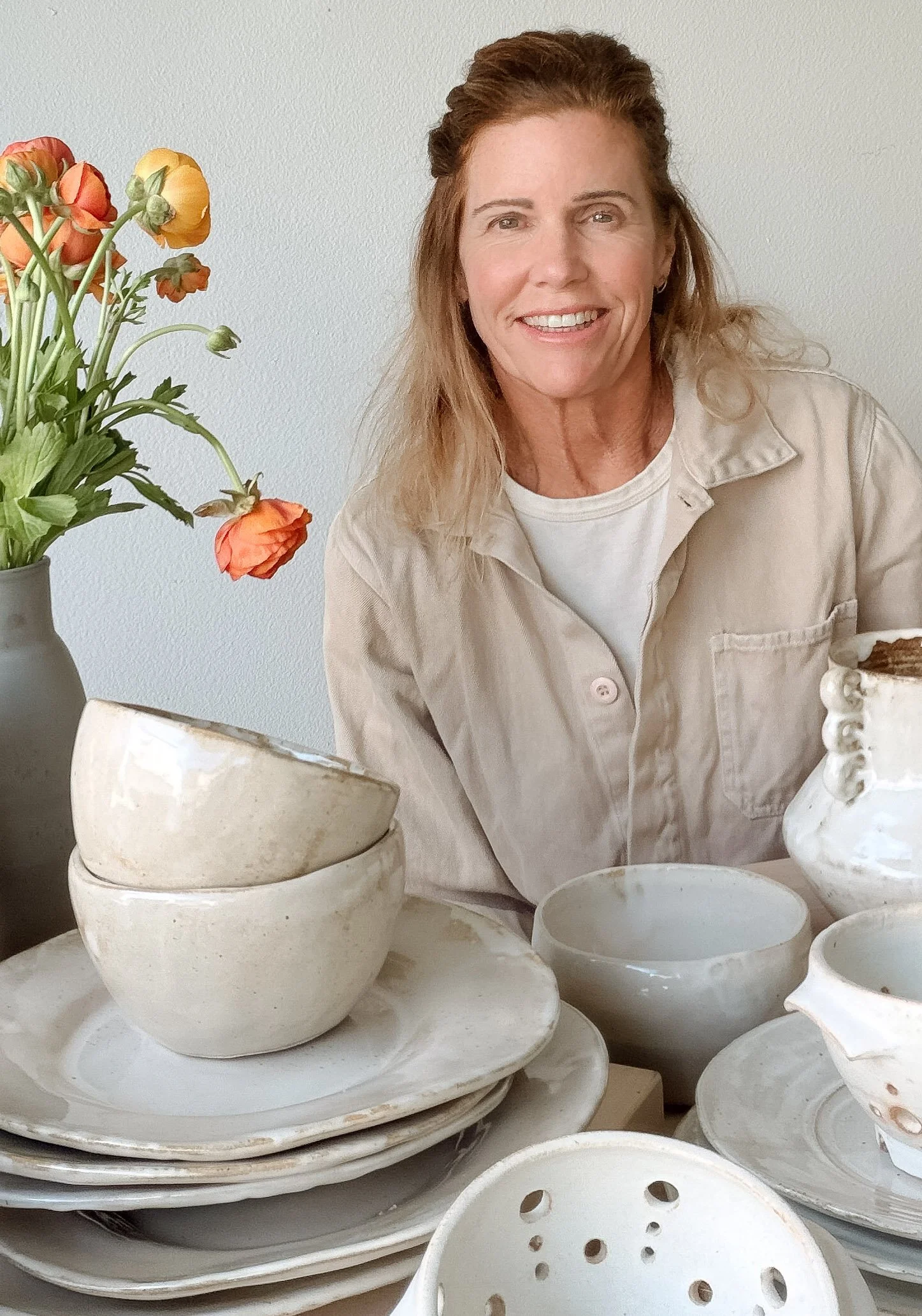 A woman with long, wavy hair smiling at a table set with ceramic bowls and plates, with a vase of orange and yellow flowers in the background.