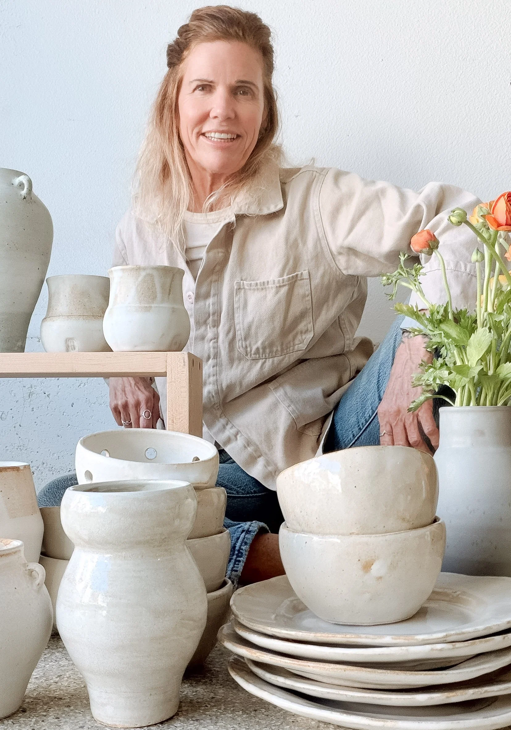 A woman with long blonde hair poses with her hand on a ceramic vase, surrounded by various white ceramic bowls, plates, and vases, with a bouquet of orange and pink flowers in a white vase.