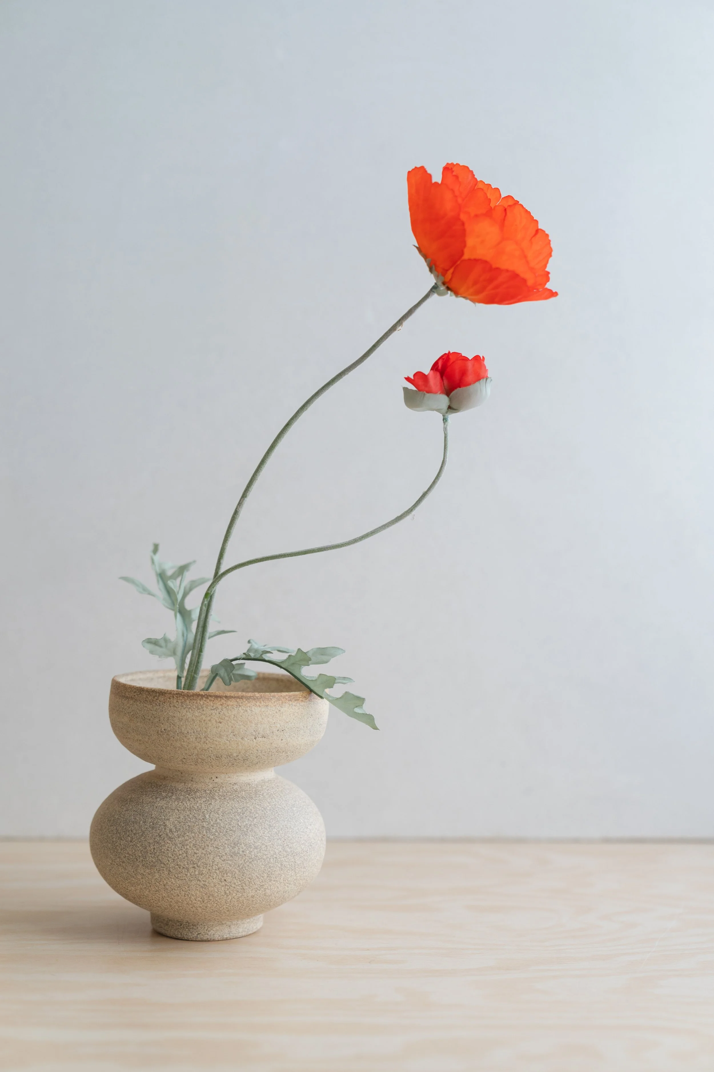 A minimalist ceramic vase with a textured surface holding two artificial poppy flowers, one red and one pink, against a plain light gray background.