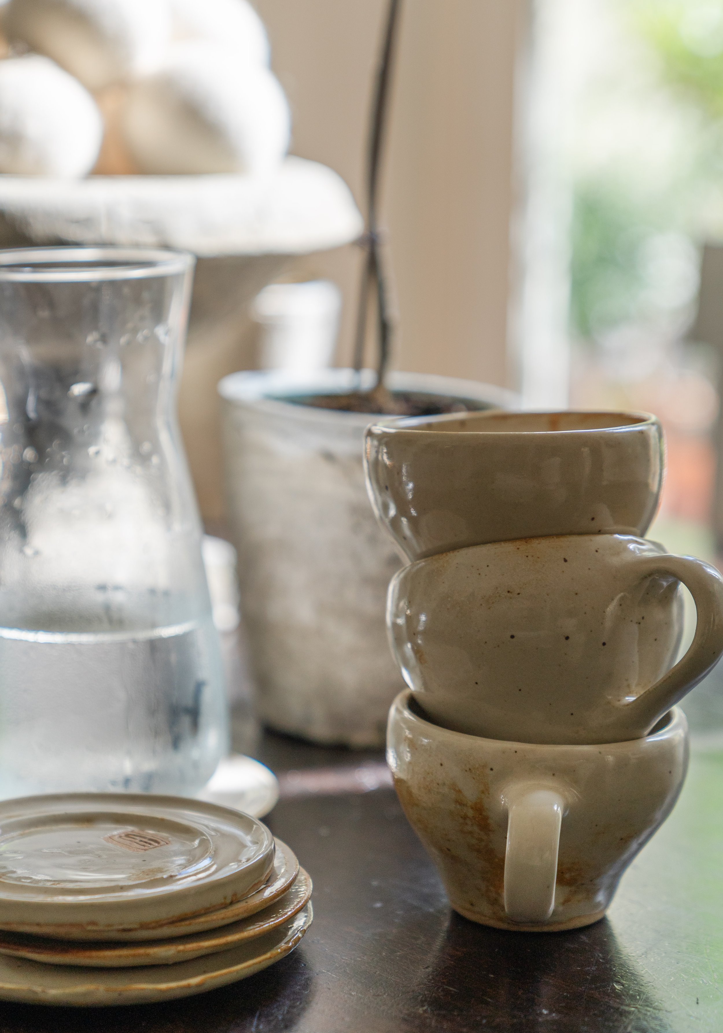 Stacked beige ceramic cups, a glass pitcher of water with condensation, and small ceramic plates on a dark table near a window with blurred green and light background.