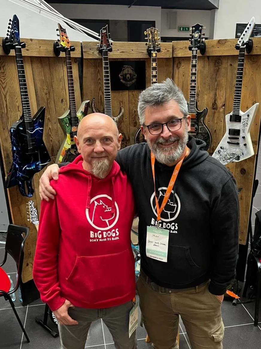 Two men, one wearing a red hoodie and the other a black hoodie, stand side by side with their arms around each other, smiling at the camera. Behind them is a display of electric guitars mounted on a wooden wall.