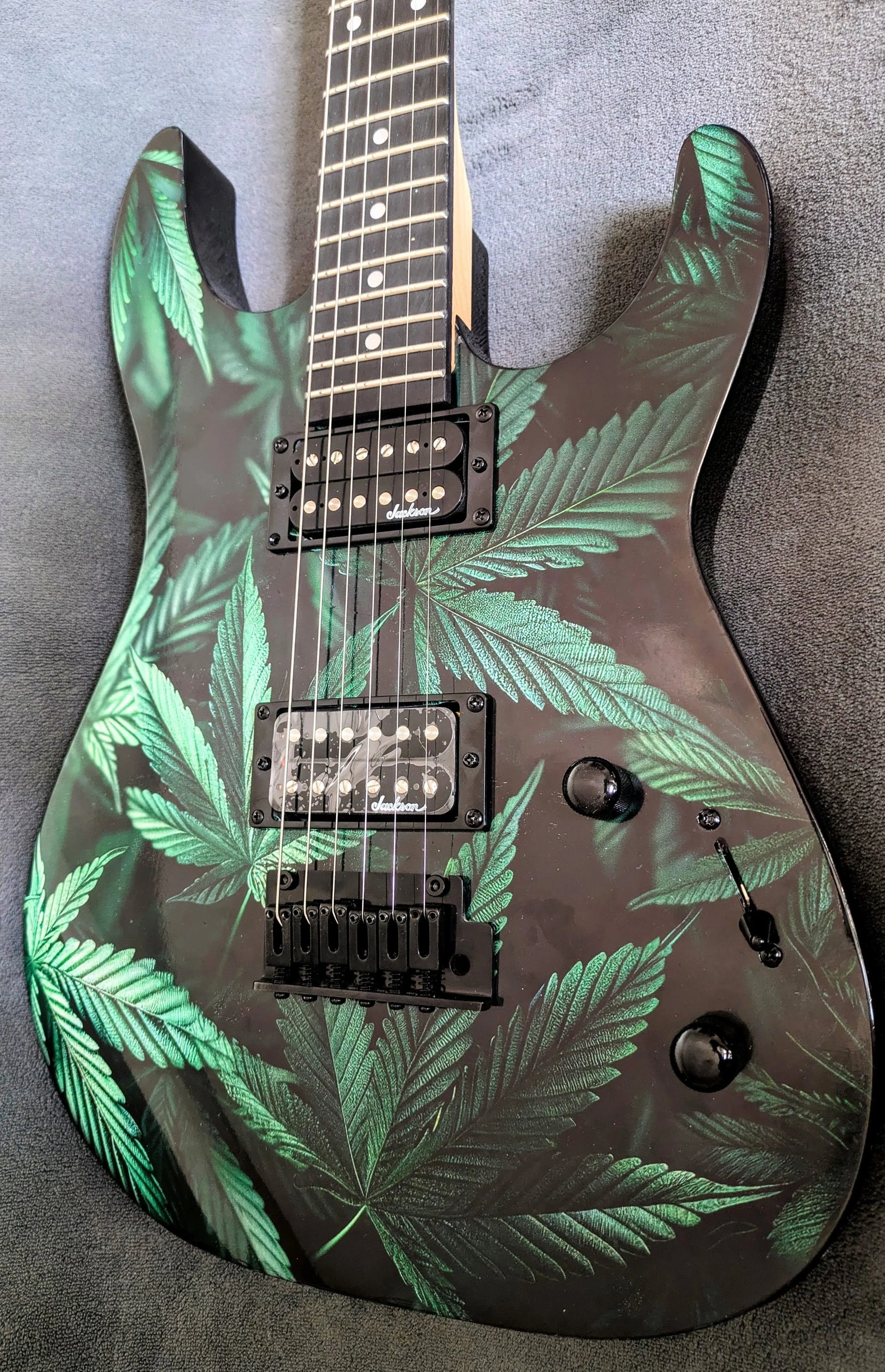 Electric guitar with a black body decorated with green cannabis leaf patterns, featuring humbucking pickups, a black bridge, and black control knobs placed on a dark surface.