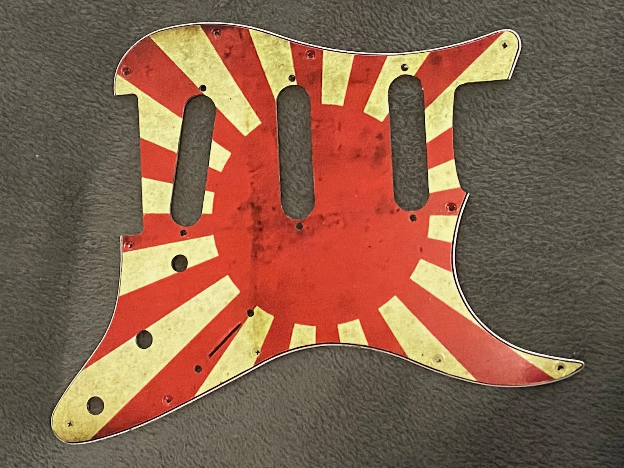 A vintage metal guitar pick guard with a red and yellow striped pattern, showing some rust and scratches, on a black textured surface.