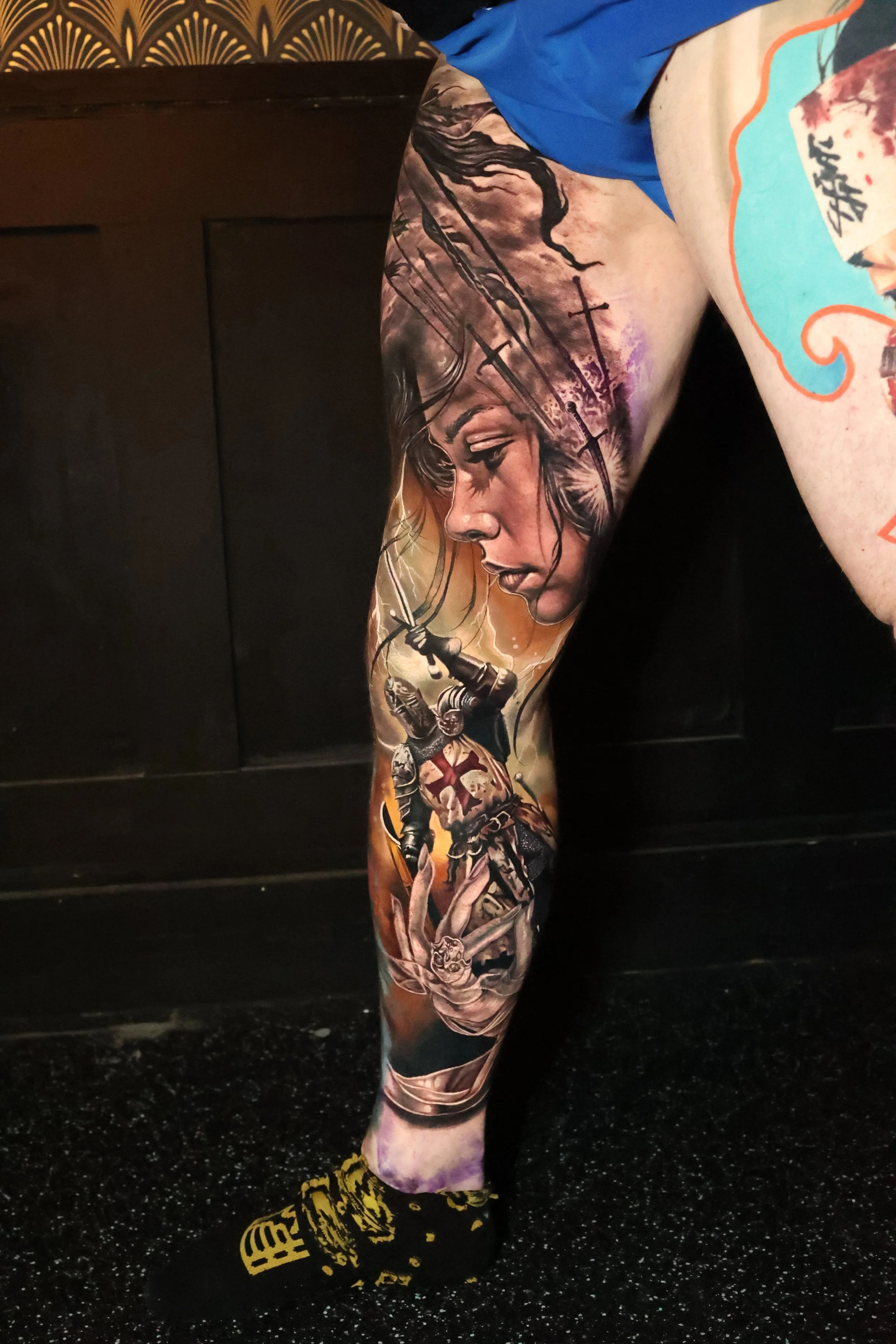 A person's leg with a colorful tattoo sleeve featuring a woman's face with black hair and a cross, along with various detailed designs. The person is wearing black and yellow boots and blue shorts, standing on a black, speckled floor.