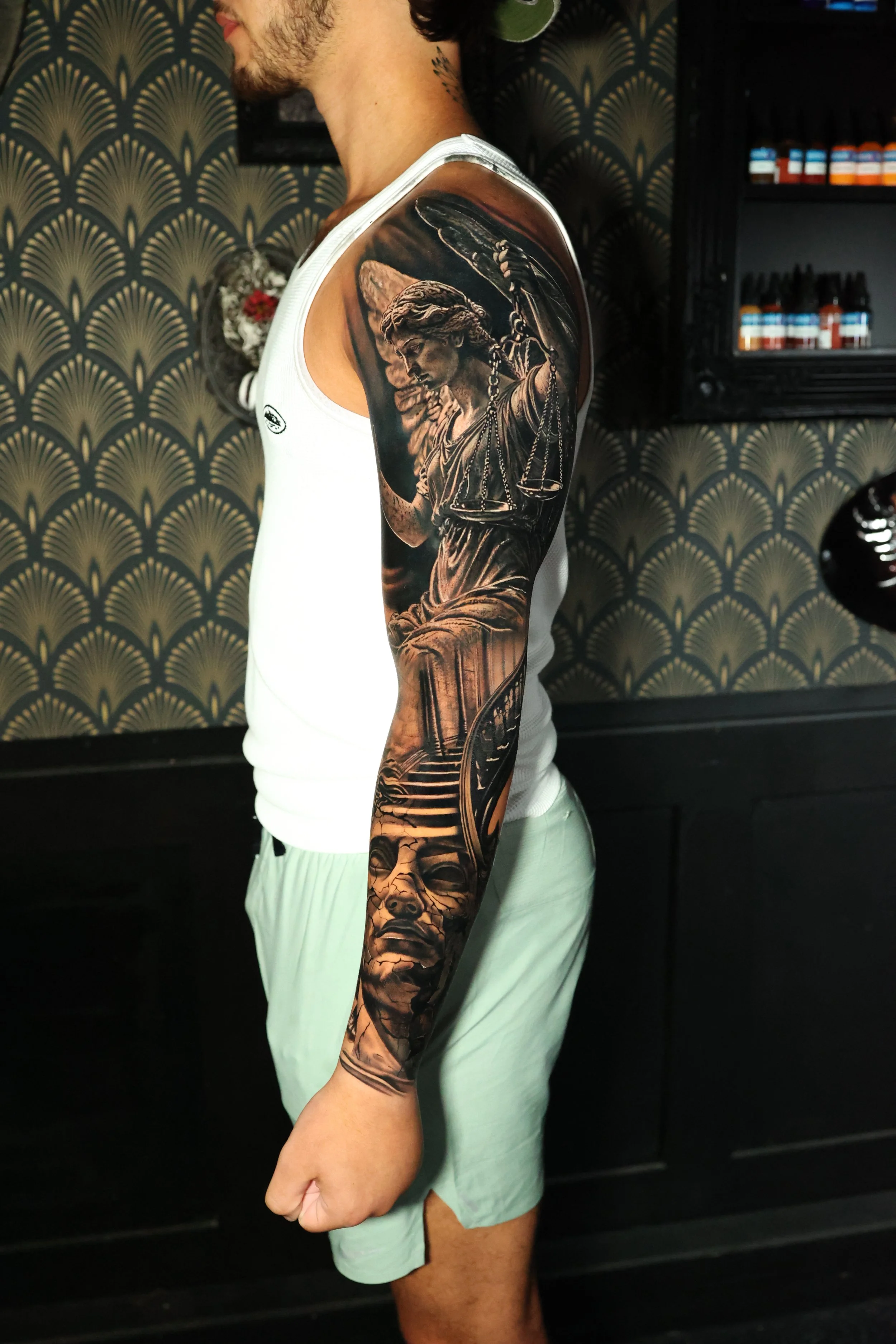 A person with a large detailed black and gray tattoo sleeve on their left arm, featuring a lady holding a set of scales and other motifs, wearing a white tank top and light-colored shorts, standing indoors with a patterned wall and shelves with bottl