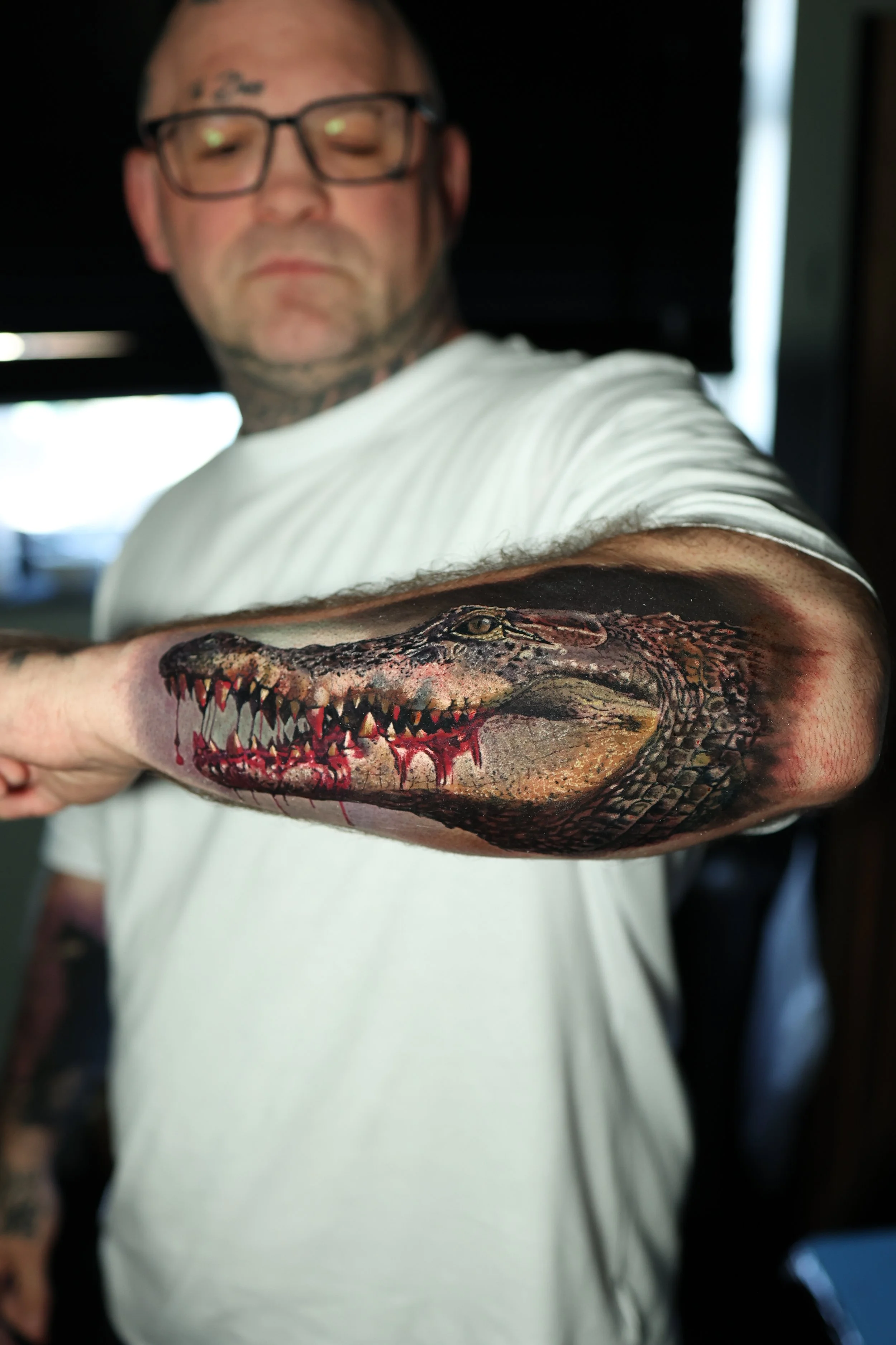 A man with tattoos on his arm and tattoos on his face, wearing glasses and a white shirt, showing a detailed realistic tattoo of a crocodile on his forearm.