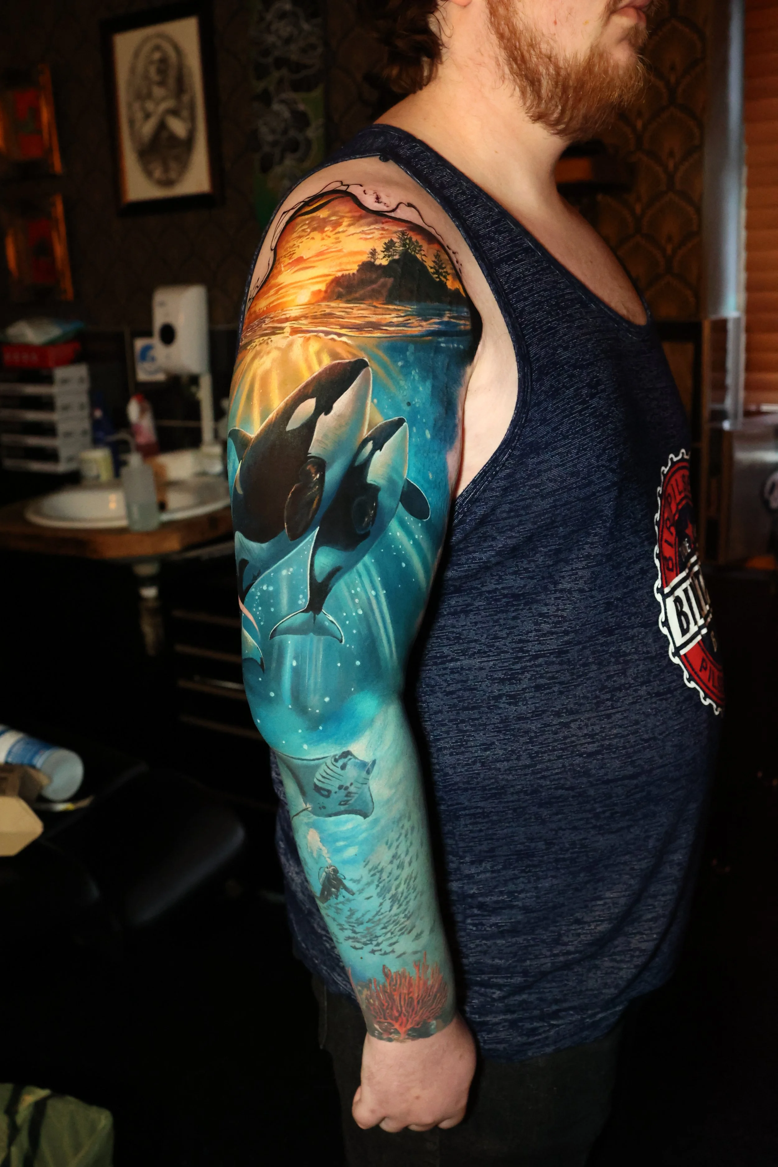 Colorful tattoo sleeve featuring a orca whale swimming in the ocean, a sunset sky, and a forested island in the background.