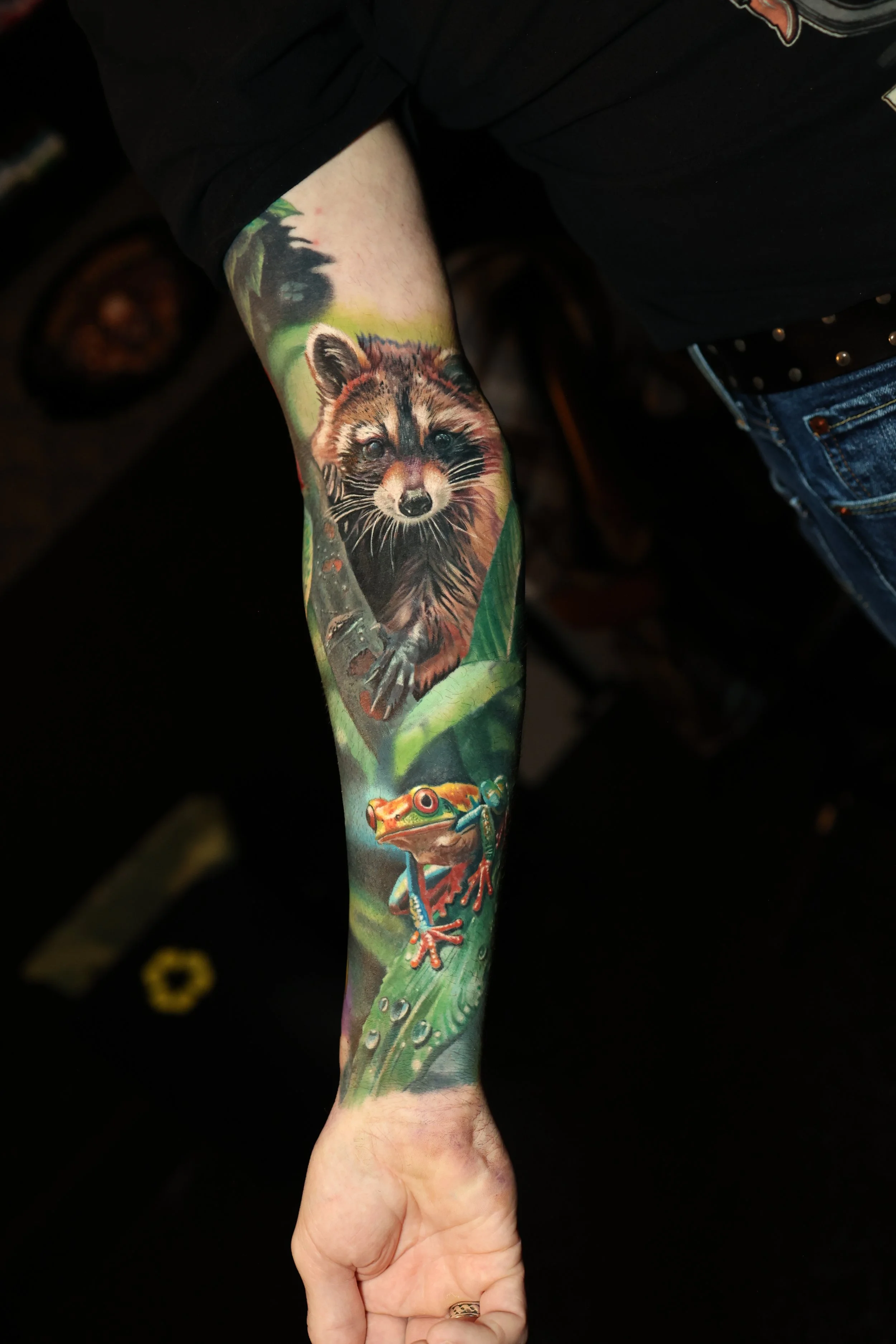 A person's arm with a detailed tattoo of a raccoon and a frog on a green leafy background.