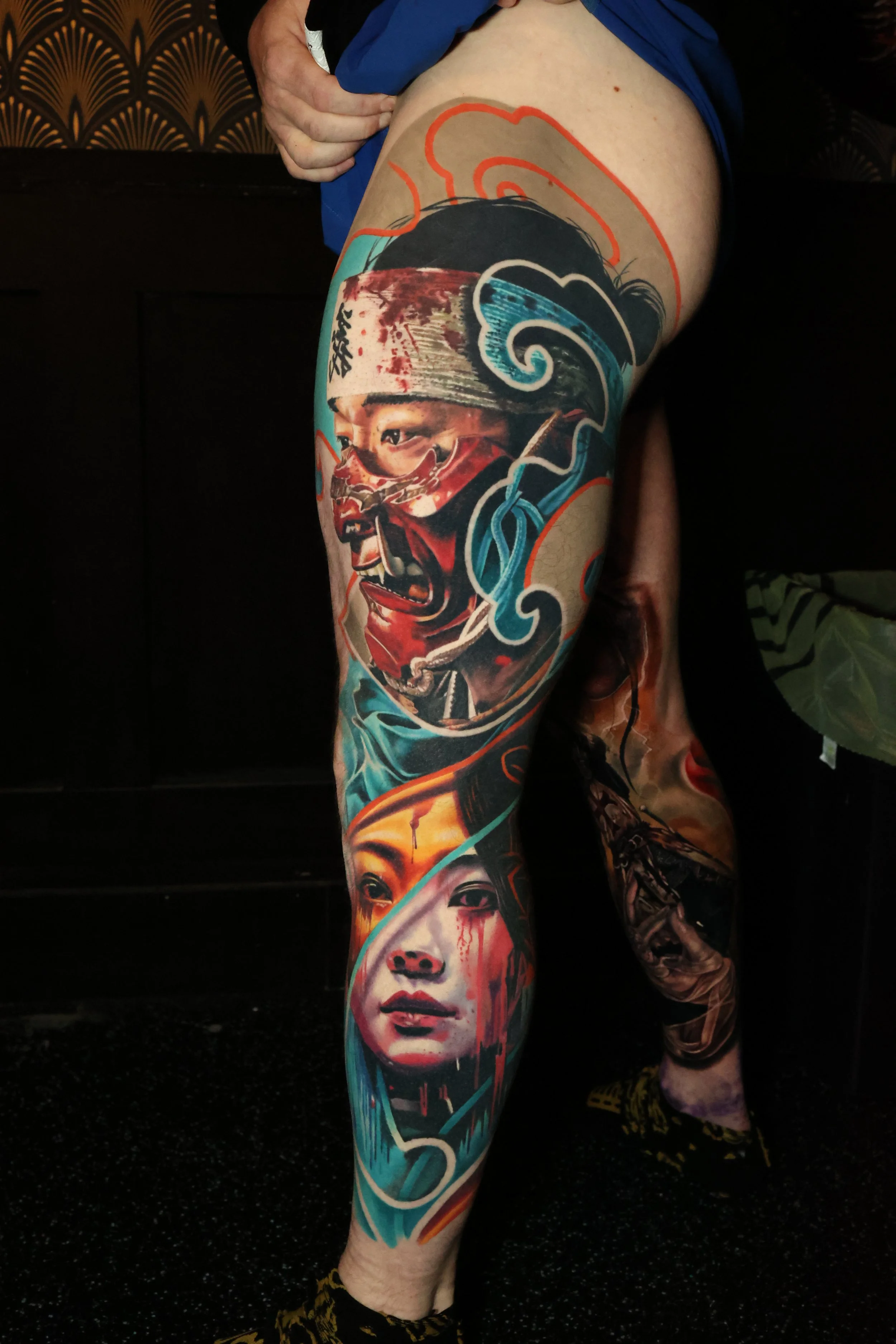 Full-leg tattoo featuring a woman's face with blood streaming from her lips, a warrior with a helmet, a cityscape, and abstract swirling patterns.