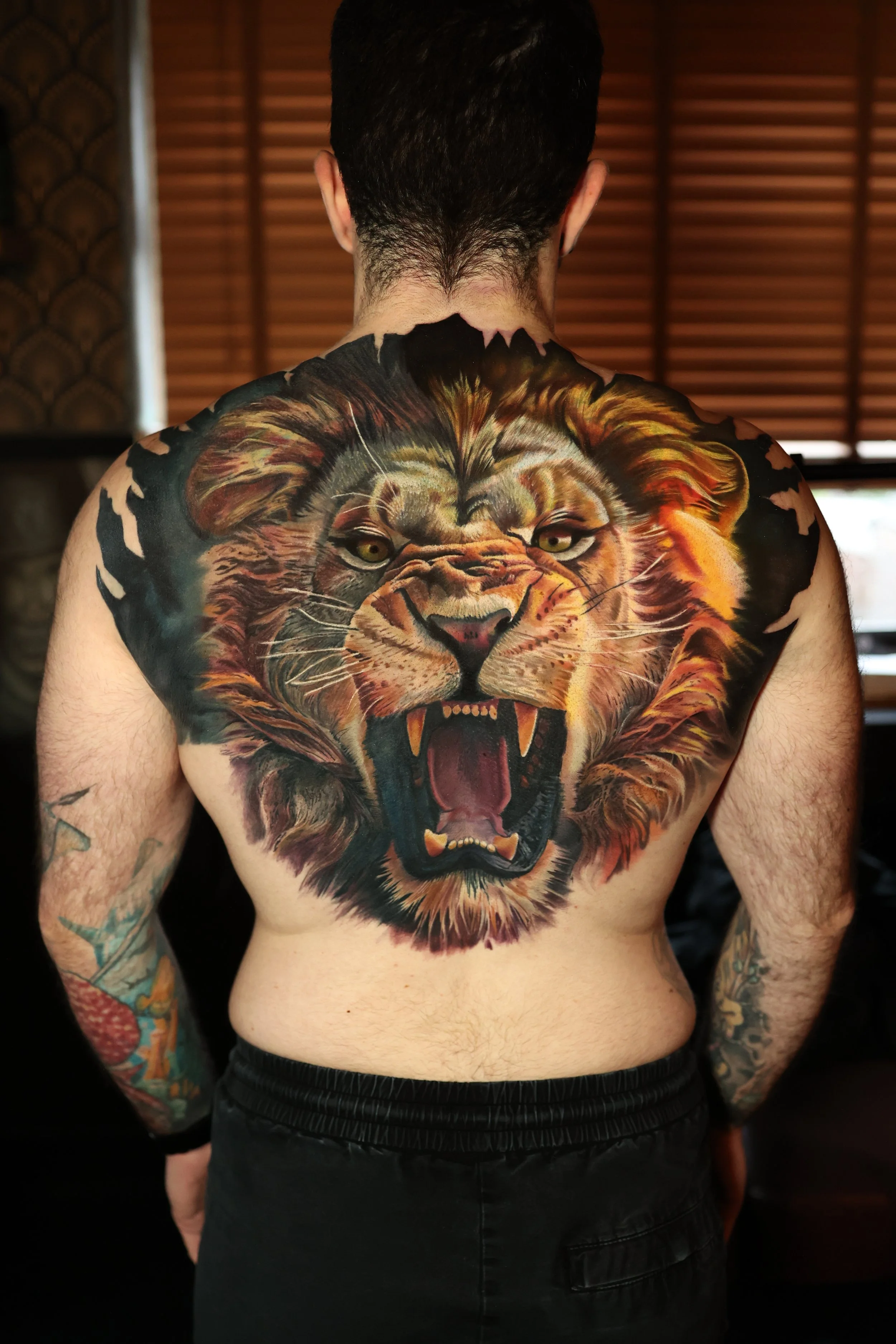 A man with tattoos on his arms and back, featuring a large colorful lion tattoo, faces away from the camera in a room with wooden blinds.