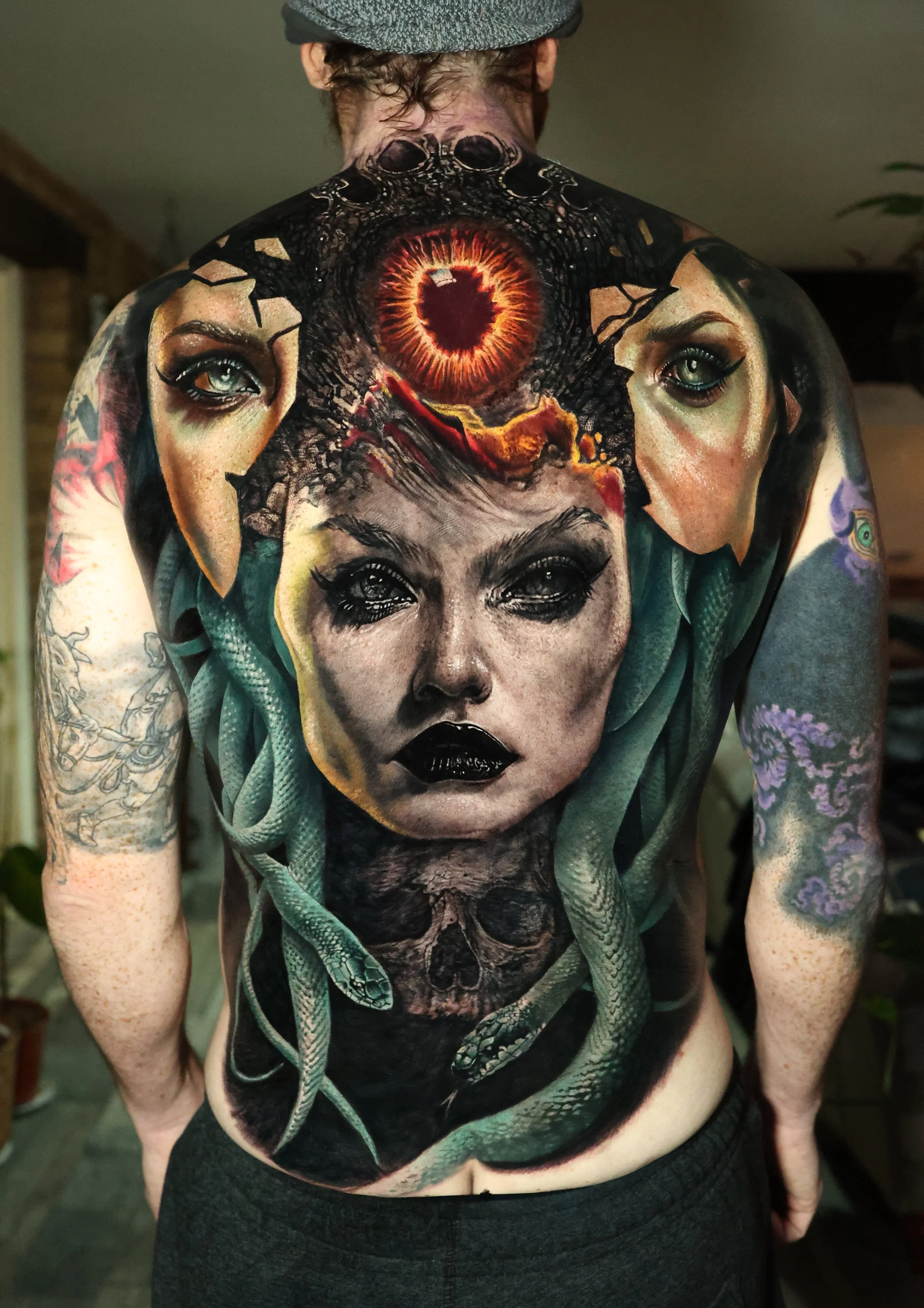 A person with a detailed tattoo on their back, featuring a large eye with a fiery heart-shaped pupil, surrounded by faces, snakes, and other intricate designs.