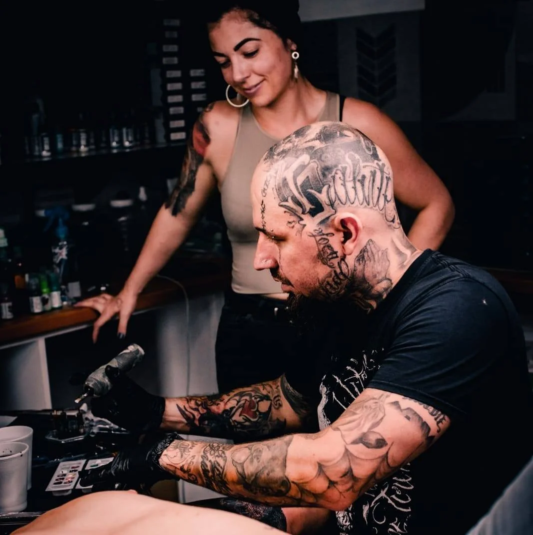 A woman with dark hair and tattoos on her arm observes a tattoo artist with many tattoos and a shaved head, working on a tattoo in a tattoo studio.