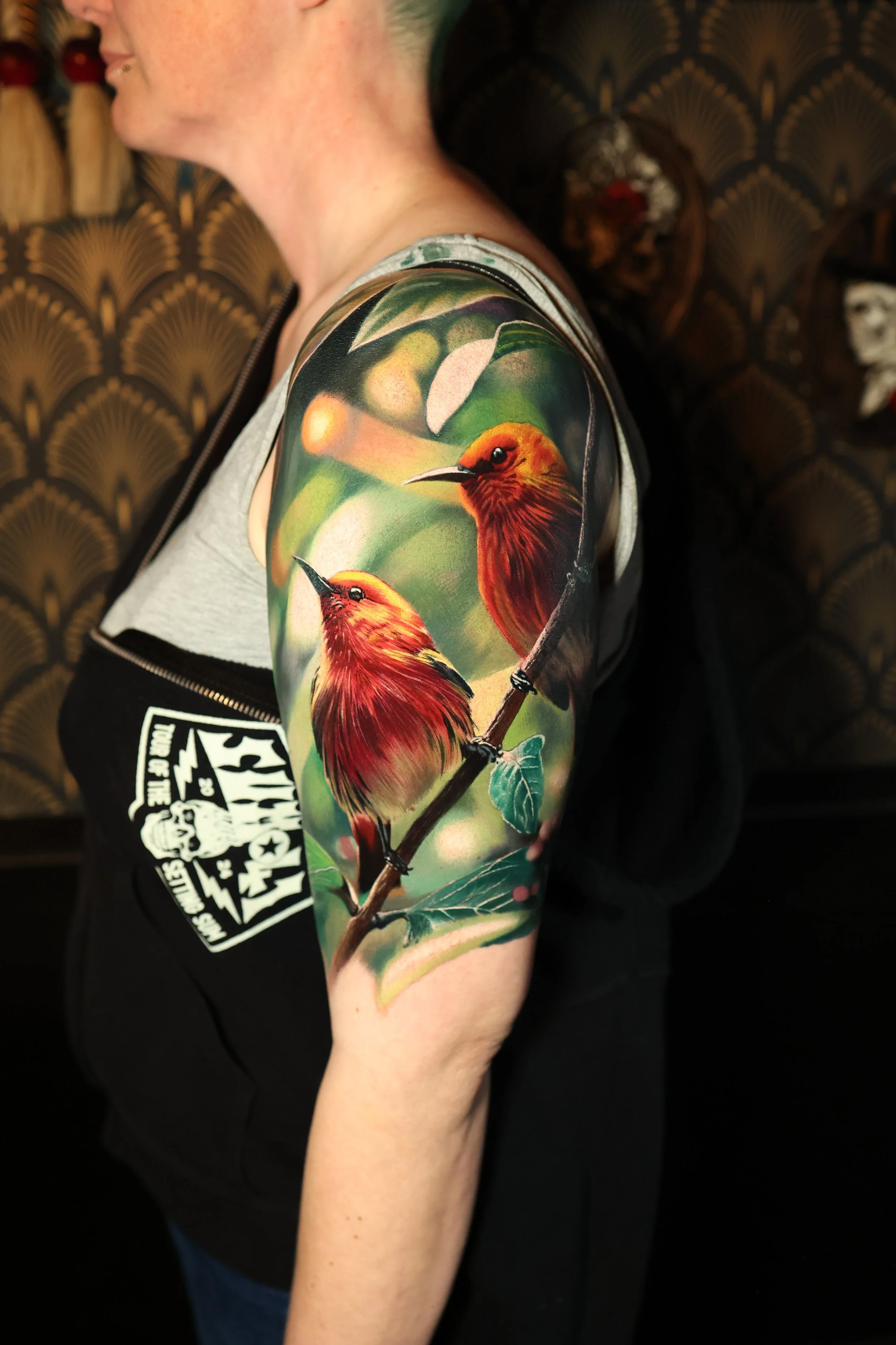 A person's upper arm with a colorful tattoo of two hummingbirds on a branch with green leaves, set against a blurred background.