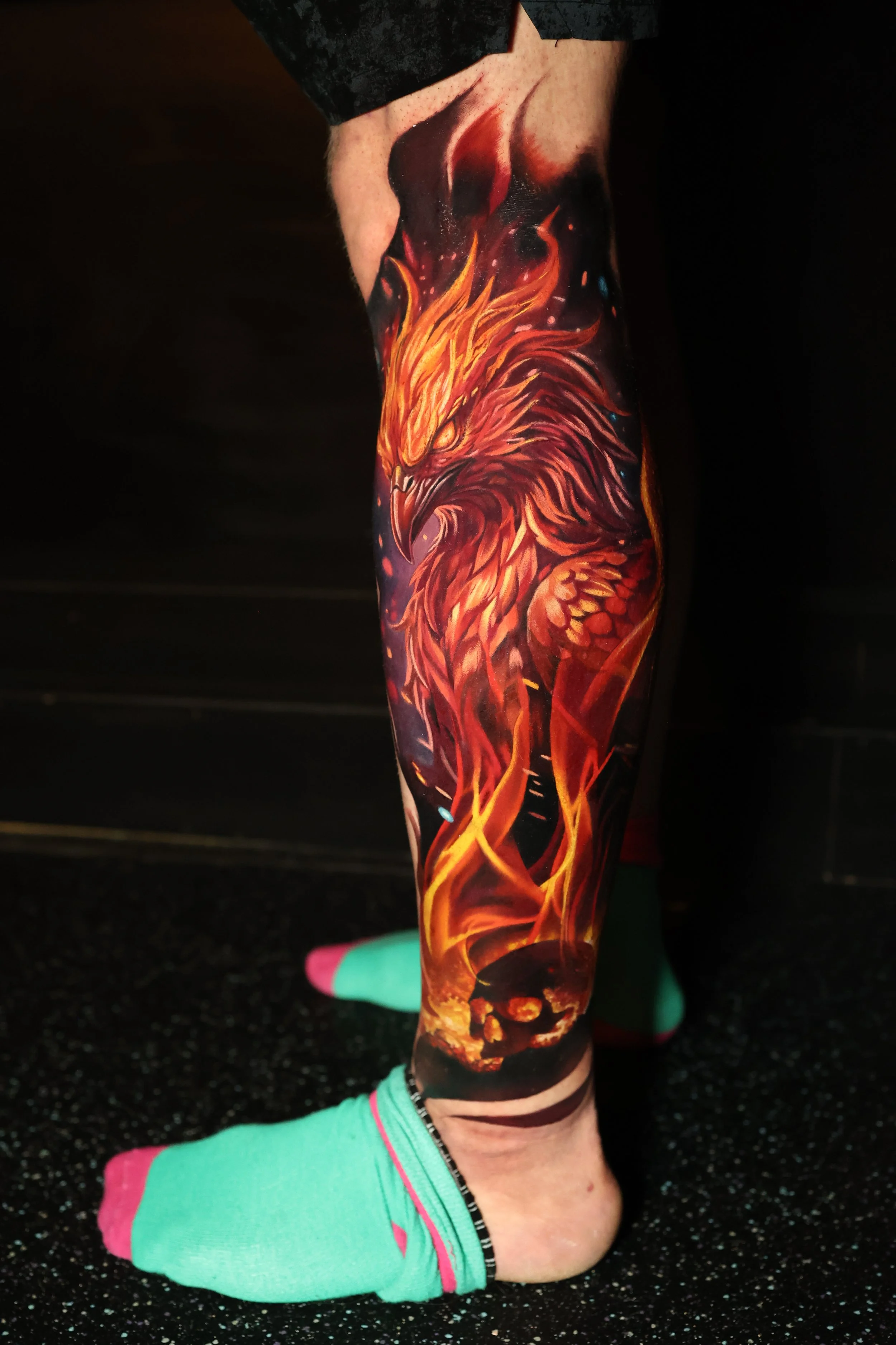 Tattoo of a fierce bird, possibly a phoenix, with bright orange and red flames, on a person's leg.