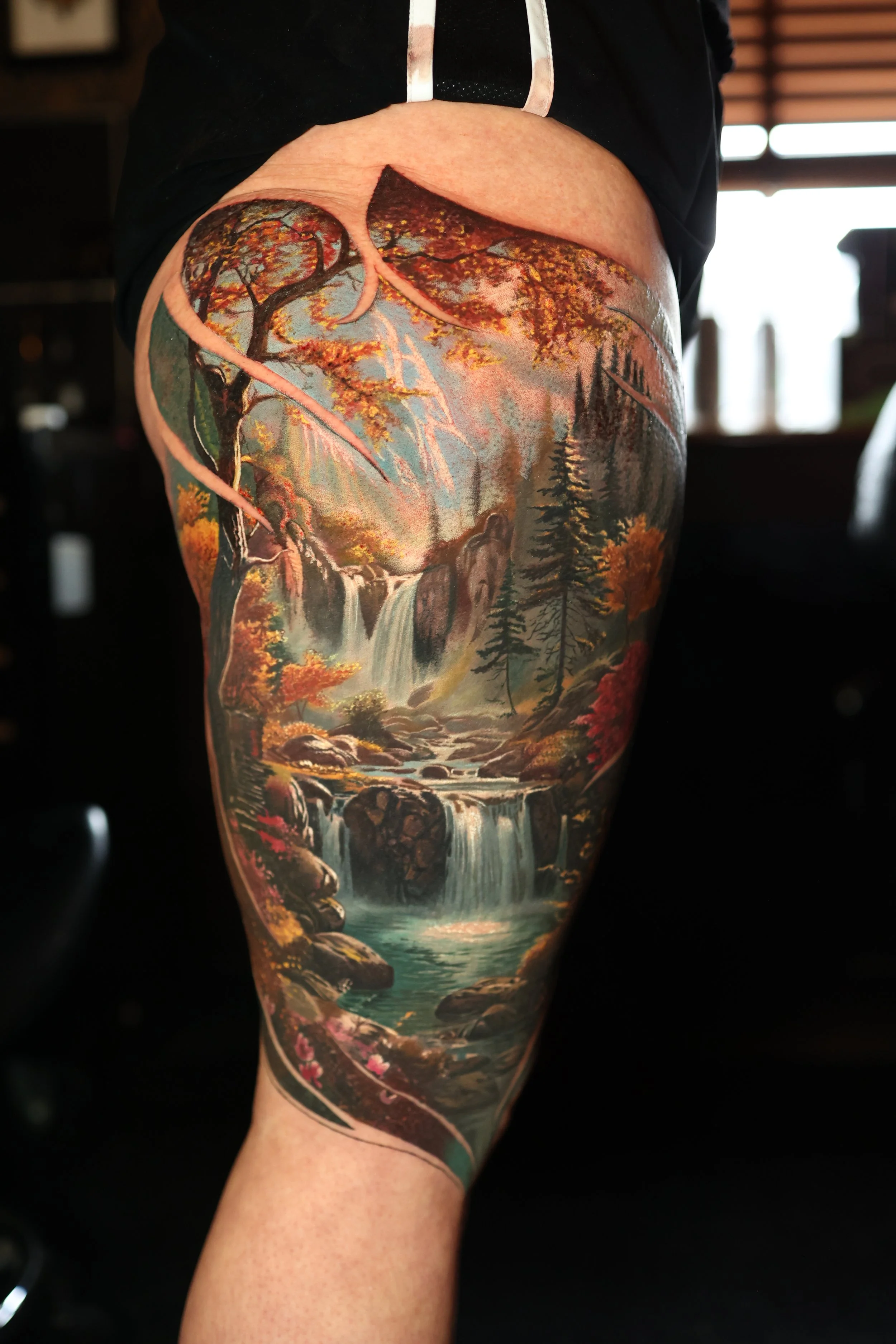 A person's leg with a detailed landscape tattoo featuring trees, a waterfall, rocky terrain, and a river in an autumn setting.