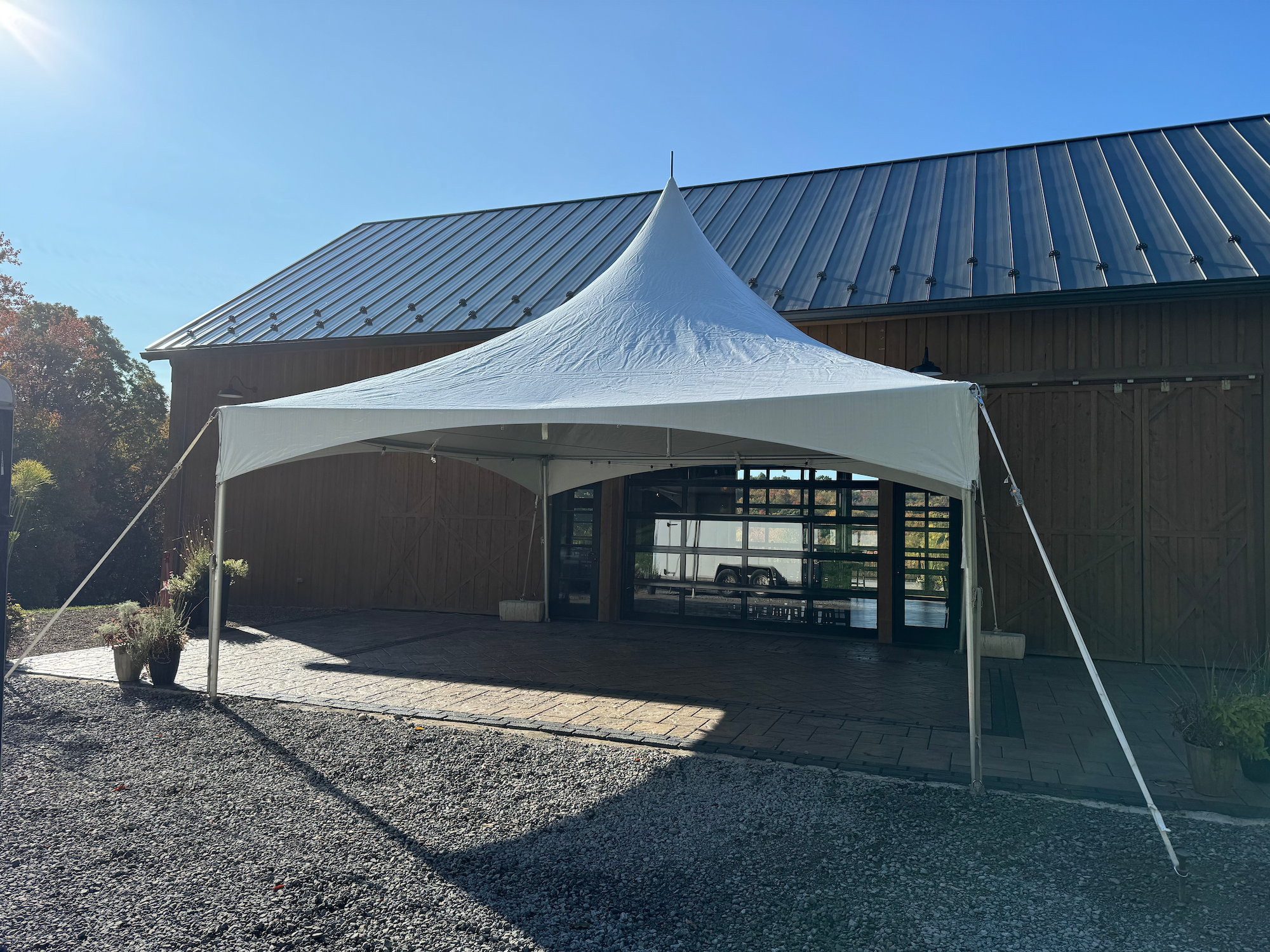 A white 20x20 High-Peak style tent in front of a barn with a metal roof, set on a brick and gravel surface, with outdoor plants and trees in the background under a clear blue sky.