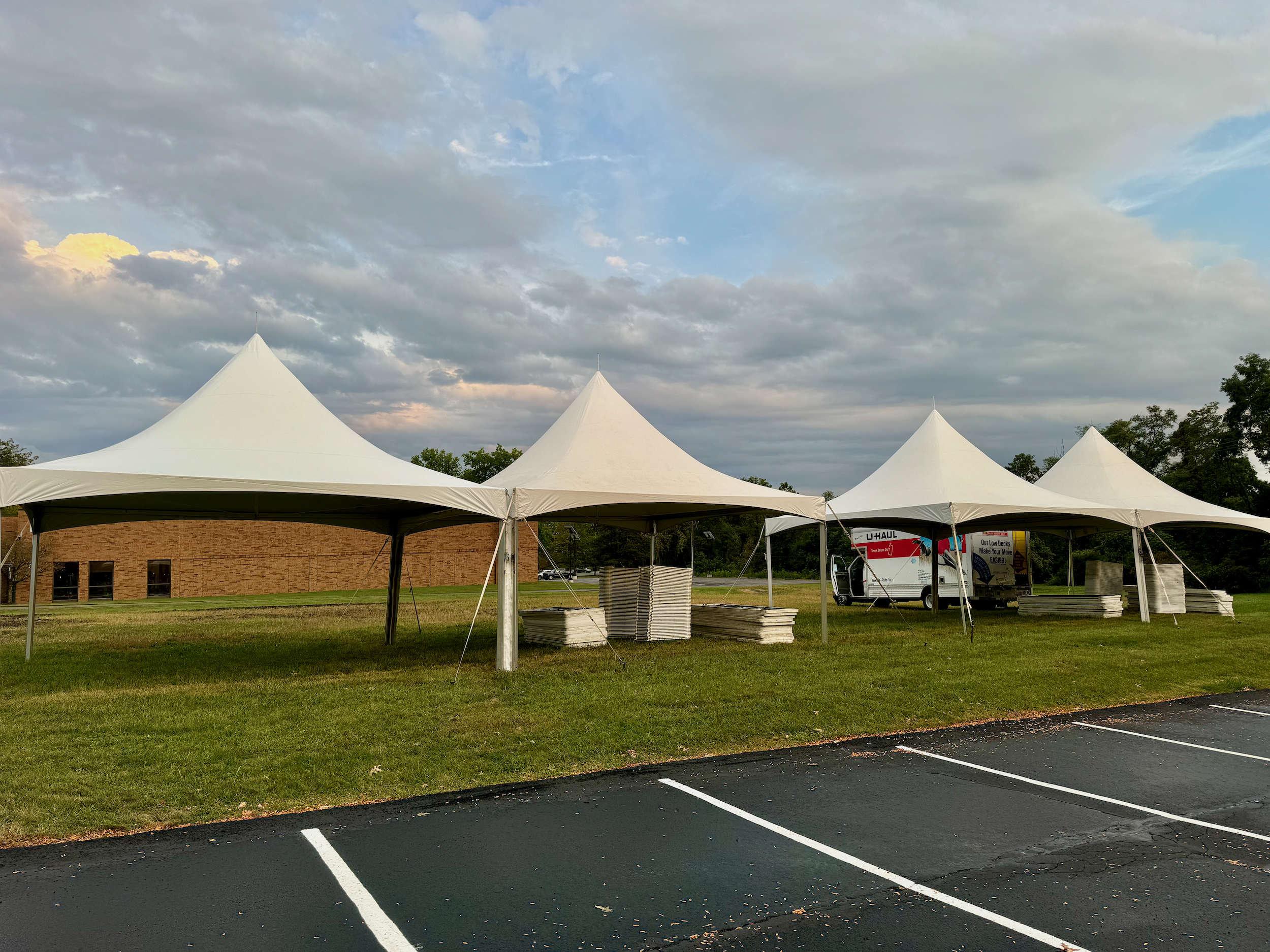 An empty outdoor parking lot with white two 20x40 high-peak tents being set up, under a partly cloudy sky.
