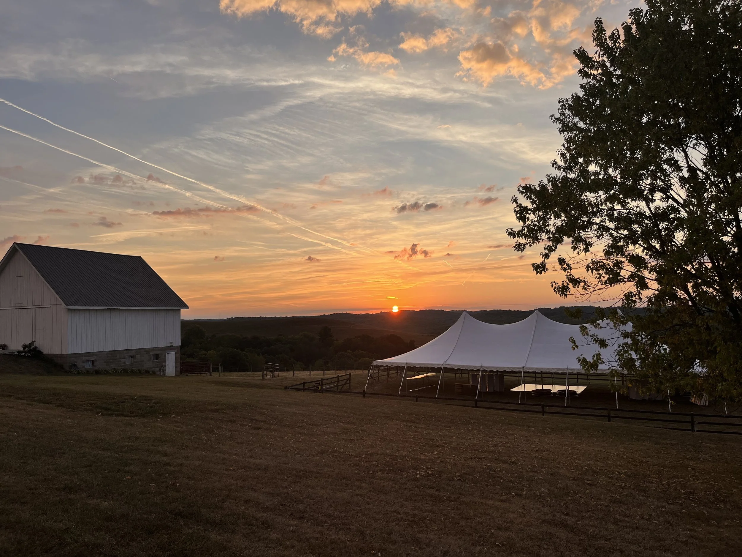 Sunset over a rural landscape with a barn on the left, a large 40x80 white wedding tent on the right, and a tree in the foreground, with streaked clouds in the sky.