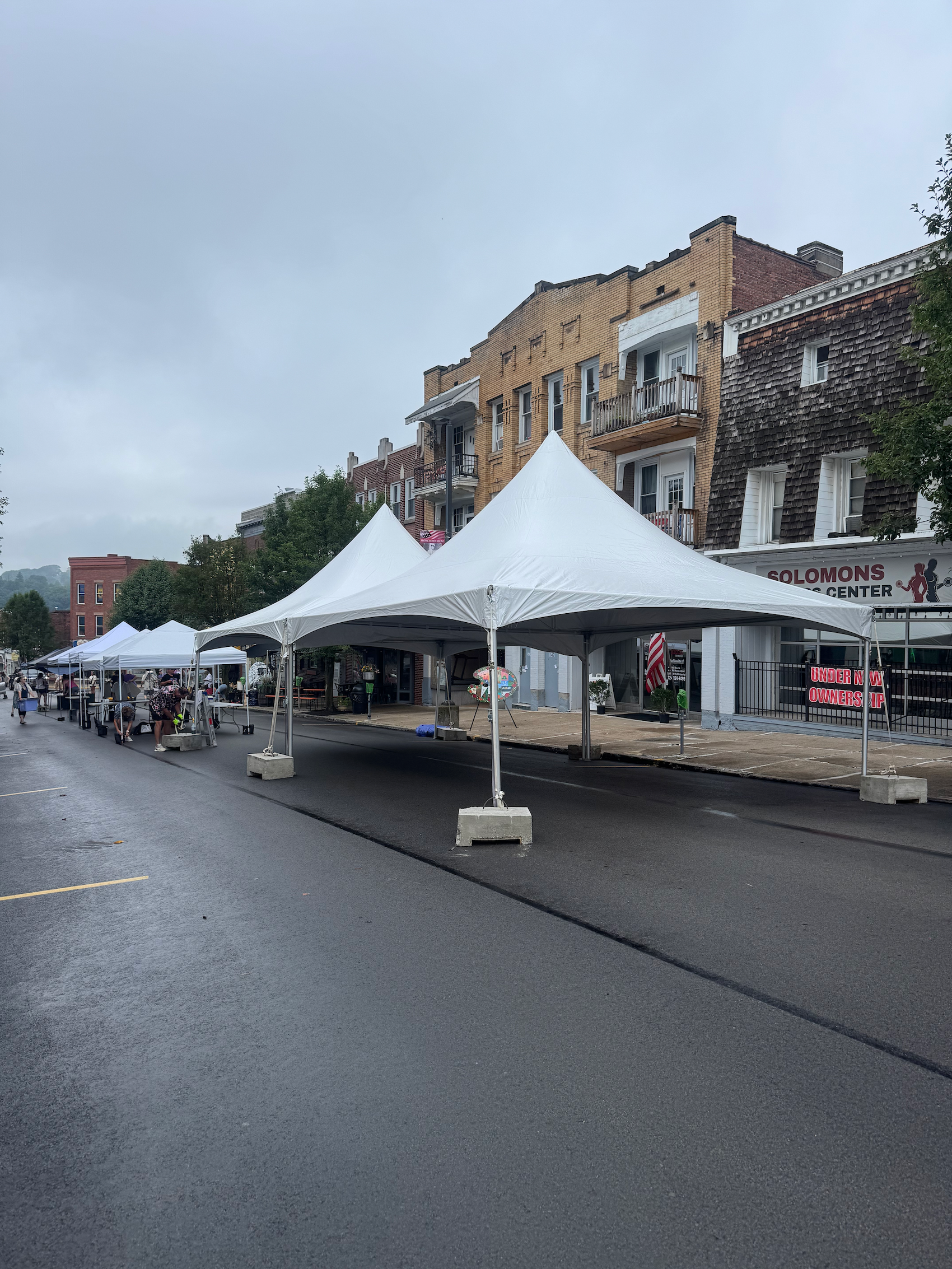 Empty street with white tents set up along the sidewalk, buildings in the background, and some people and items visible near the tents.