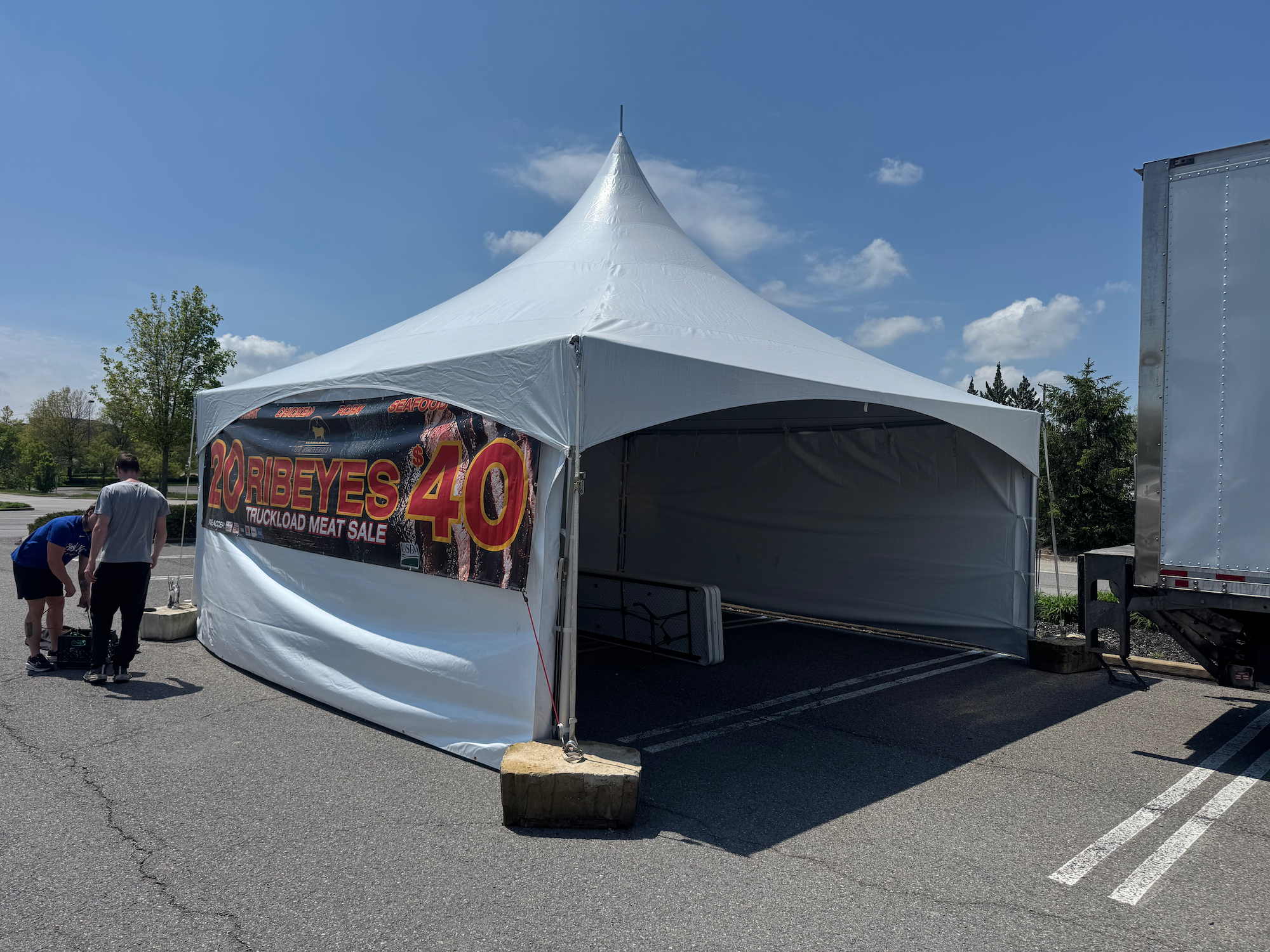 Empty white tent in parking lot with a banner advertising meat sale, mentioning 20 ribeyes for $40, with blue sky and a few trees in the background.