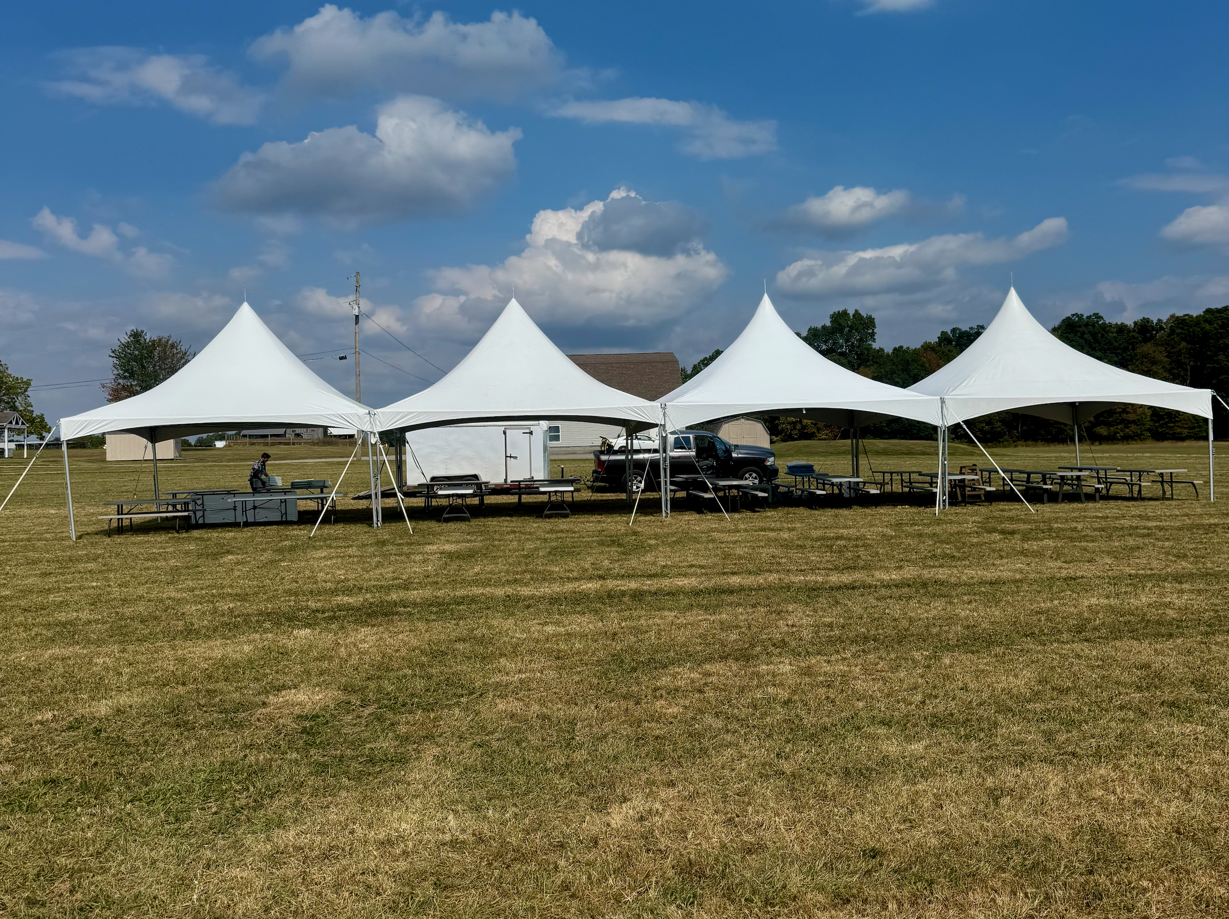 View of a large outdoor event tent with multiple peaks, set up on a grassy field under a partly cloudy sky; various tables and chairs are arranged under the tent, with a truck and trailer parked behind it.