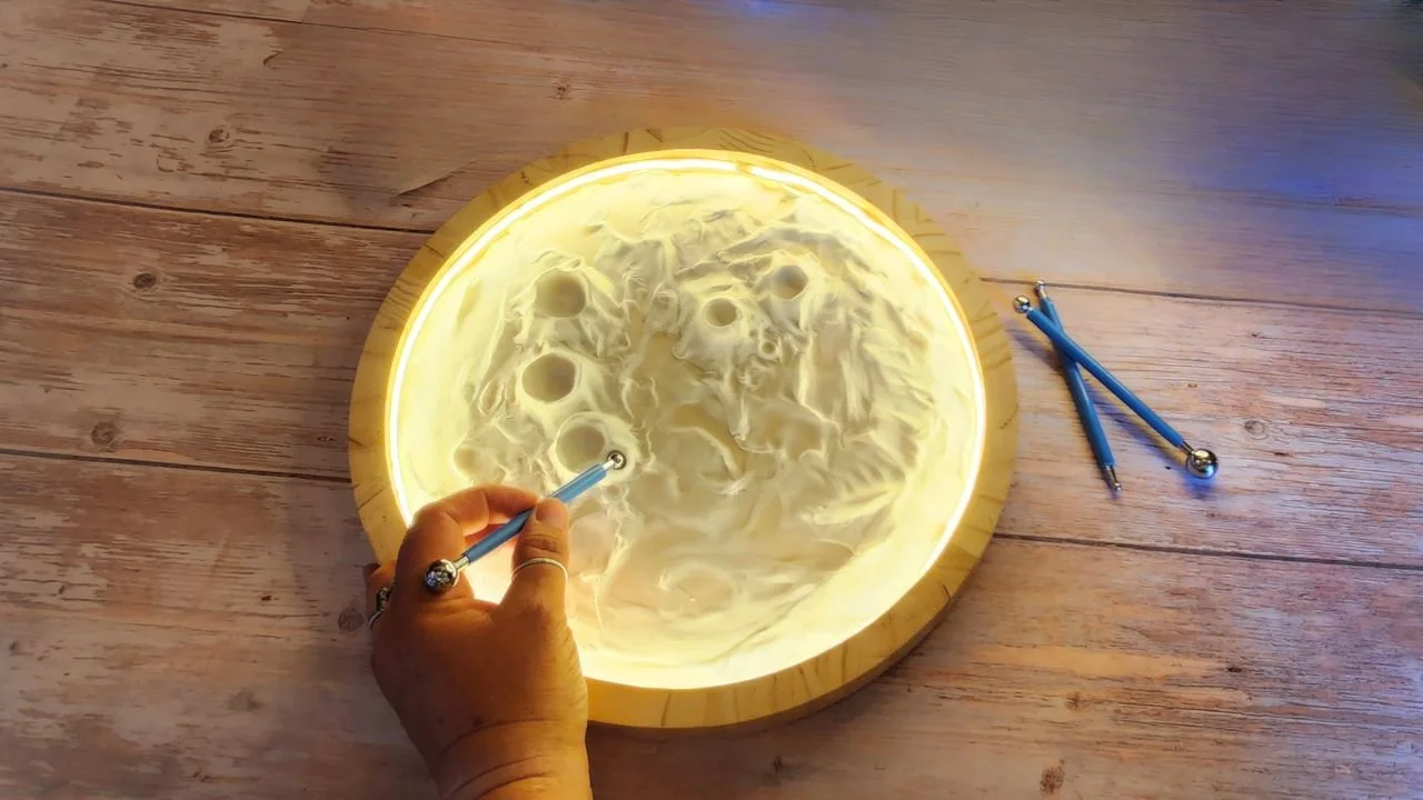 A glowy moon artwork is being created. A hand holds a stylus tool, creating craters within a round frame that is lit and looks like a moon.