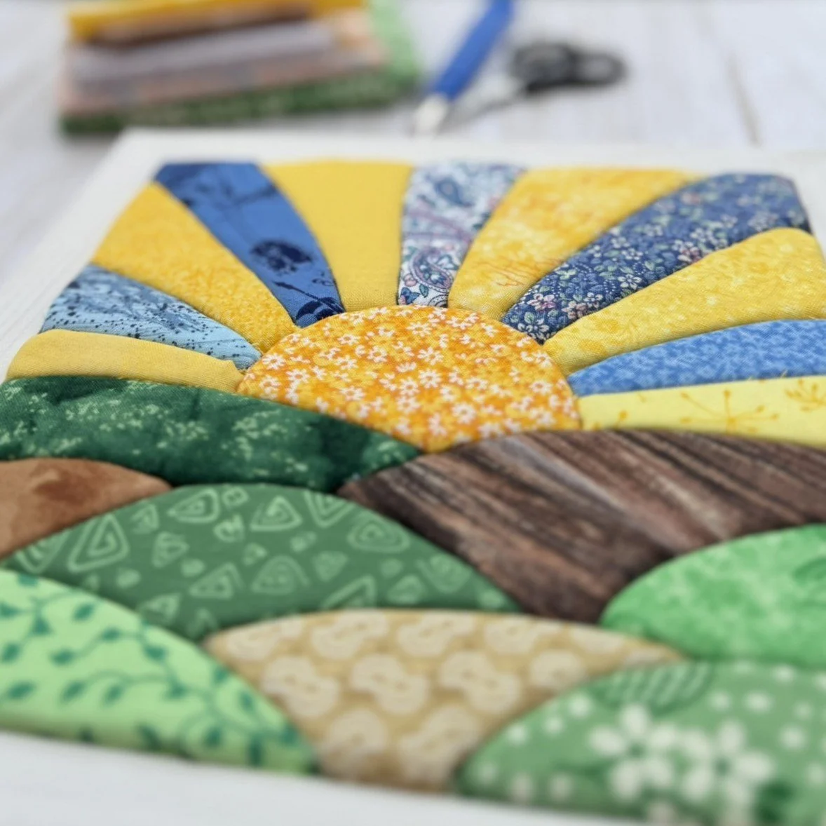 Framed kinusaiga artwork consisting of fabric pieces that make a sunrise design in yellows, blues, greens, and browns. Blurred in the background are a pair of scissors, a seam ripper, and a stack of fabric scraps.