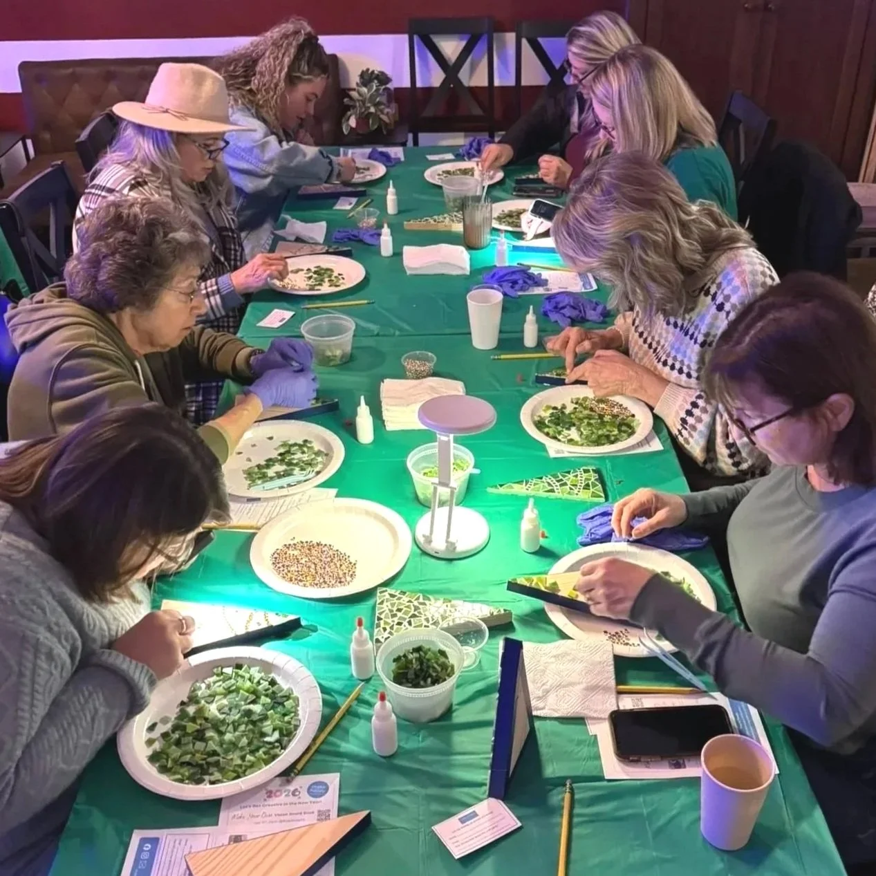 Group of women seated at a long table, engaging in a mosaic craft activity involving small green pieces of glass. There are tools and supplies spread out on the table.