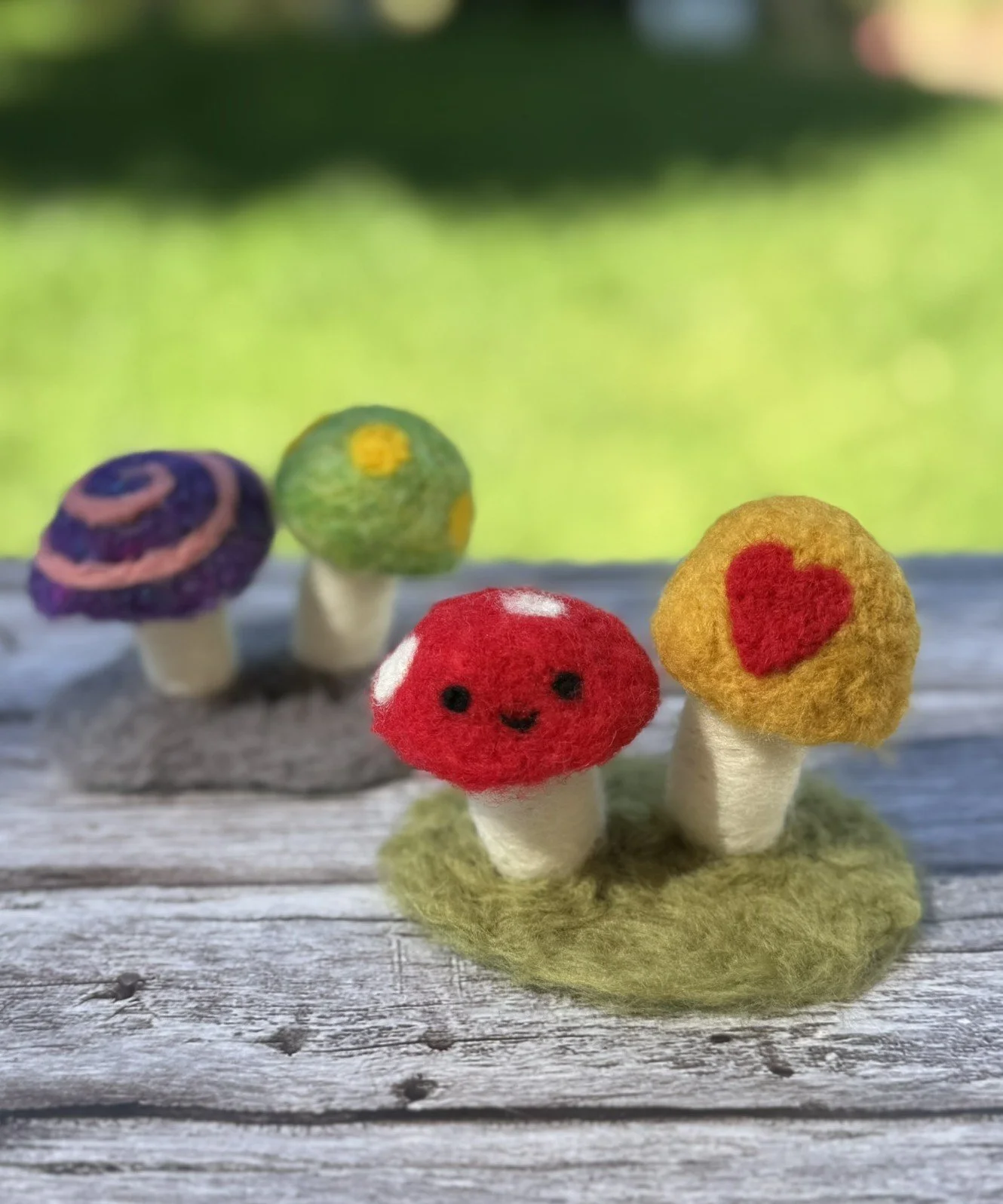 A pair of needle-felted mushrooms in front of a blurry green background. The larger mushroom has a red cap with white dots and the smaller has an orange and yellow cap. Both have cream-colored stems, and are on a green wooly base.