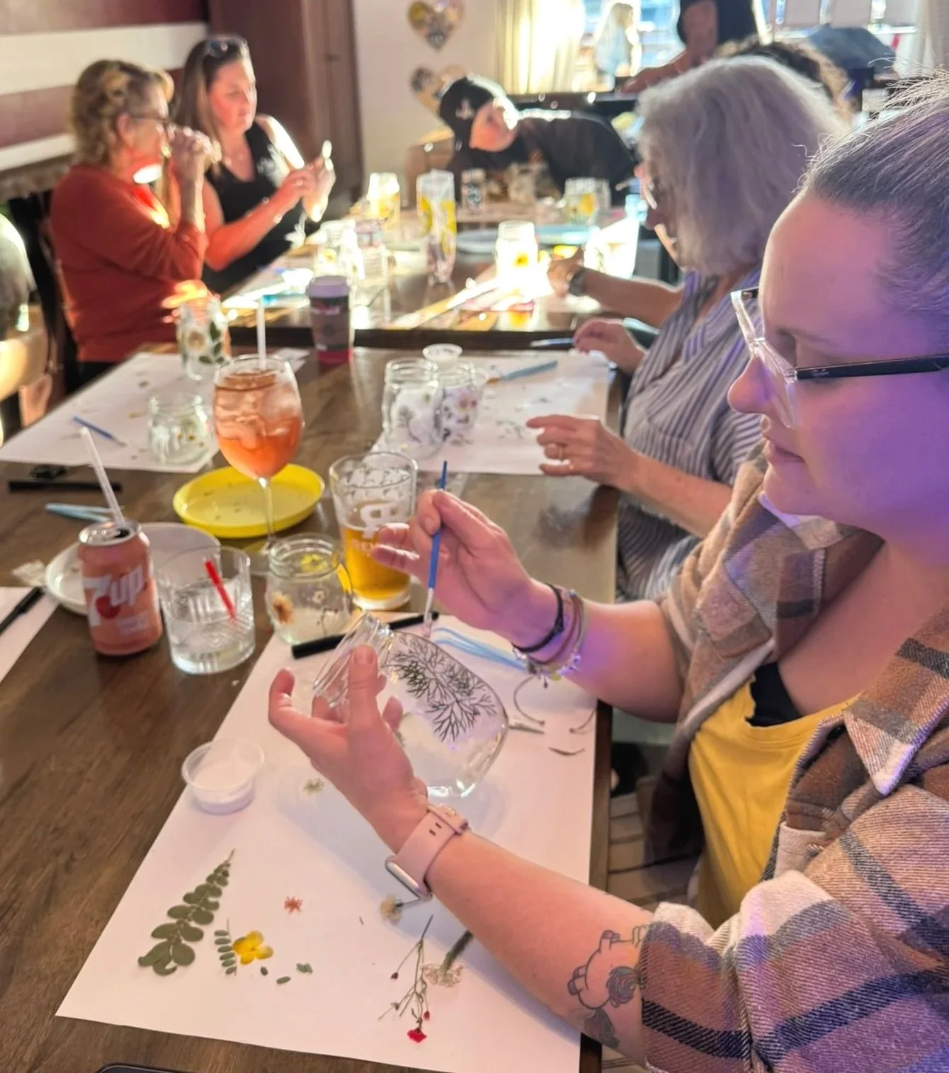 A group of people seated at a long wooden table engaging in a craft activity. Some are using brushes on glass jars, with various drinks and craft supplies scattered across the table. The background shows more participants and a cozy indoor setting with sunlight coming through windows.