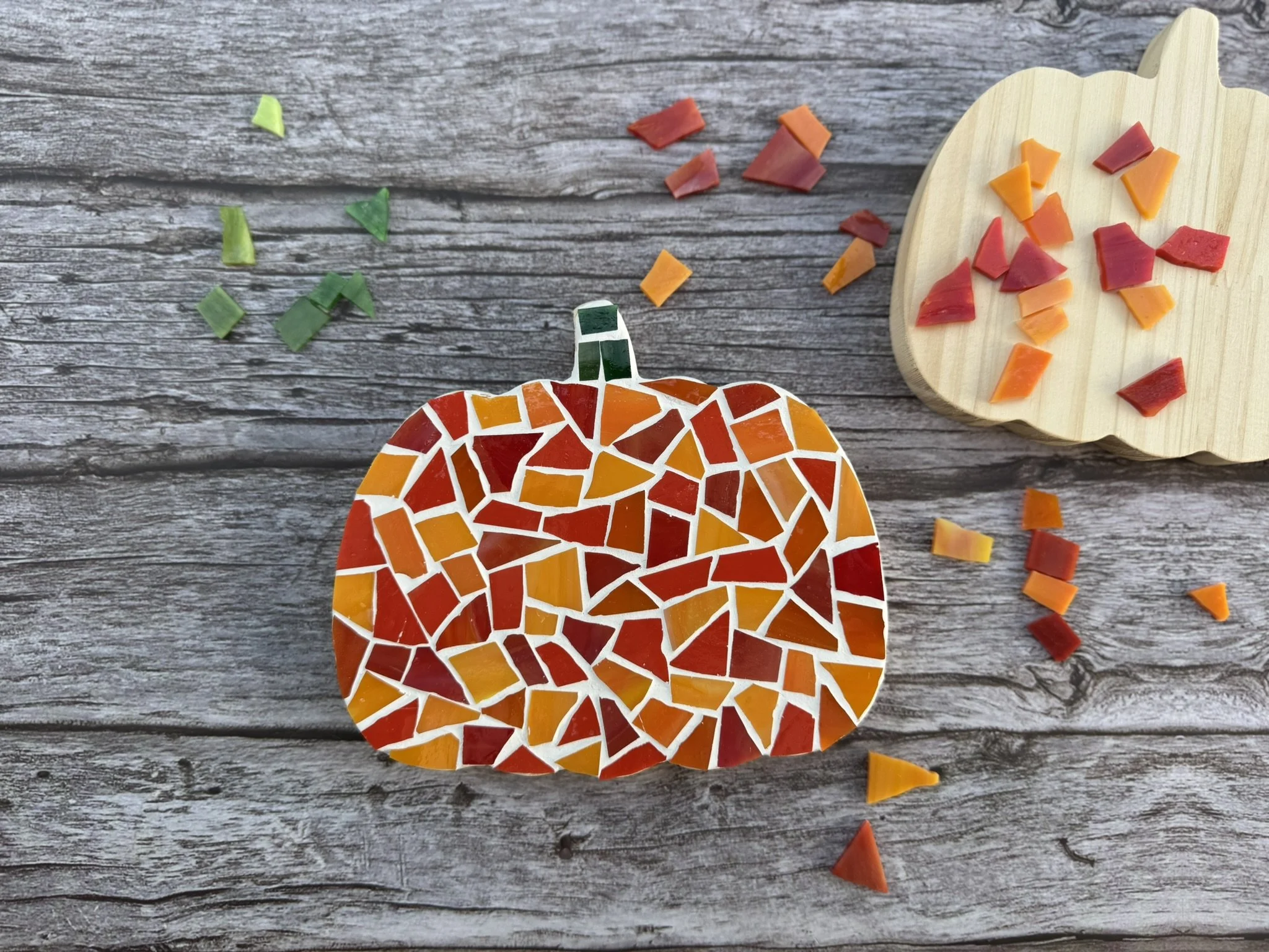 A mosaic in varying shades of orange glass is in a pumpkin shape. It's on a neutral wood background with orange and green glass pieces scattered around. 