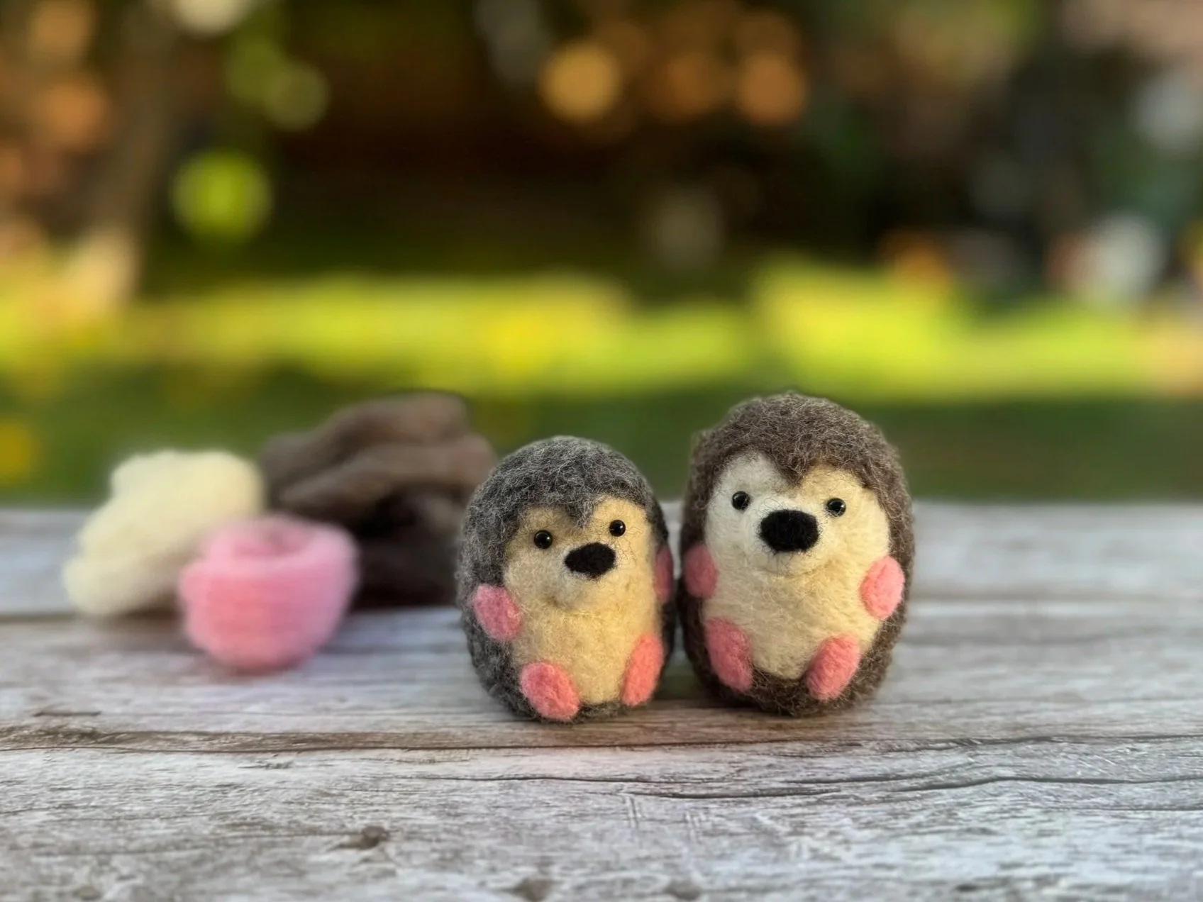 Two needle-felted hedgehogs on a wooden surface against a blurred background. There are three small puffs of felting wool behind them.