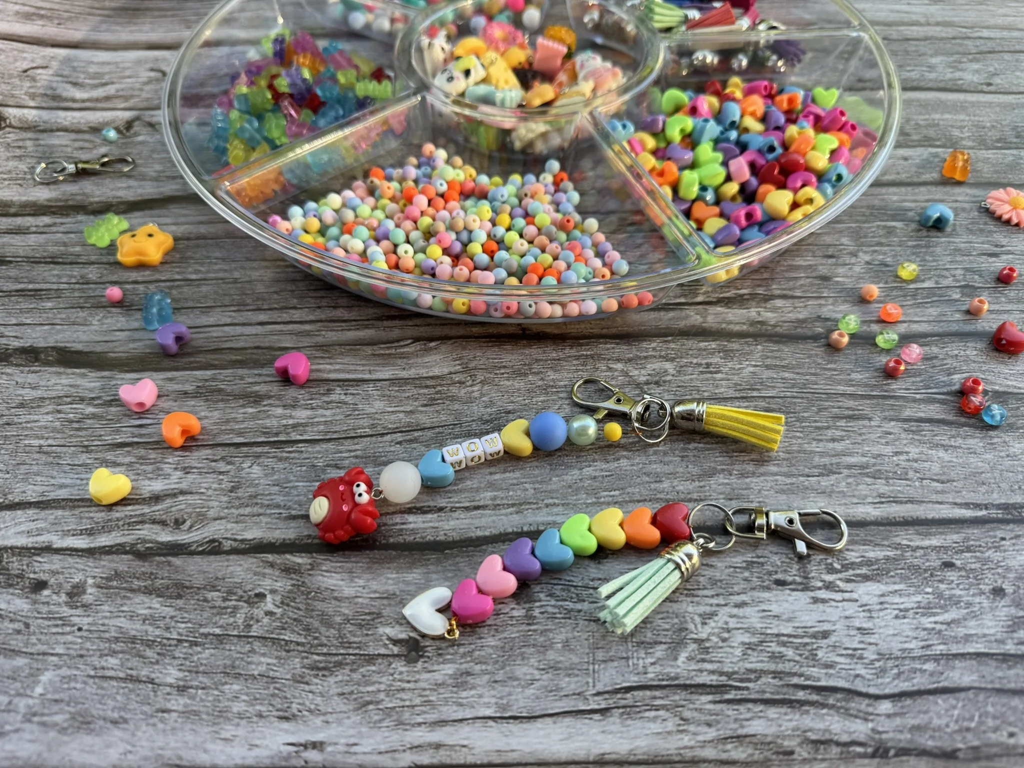 Two beaded keychains sit on a wooden surface. A tray of colorful beads is in the background, and several beads are scattered around.