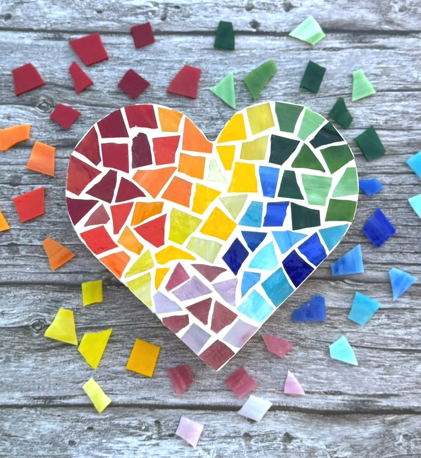 A mosaic heart consisting of rainbow-colored glass pieces. on a neutral wooden background. Additional colorful glass pieces surround the heart.