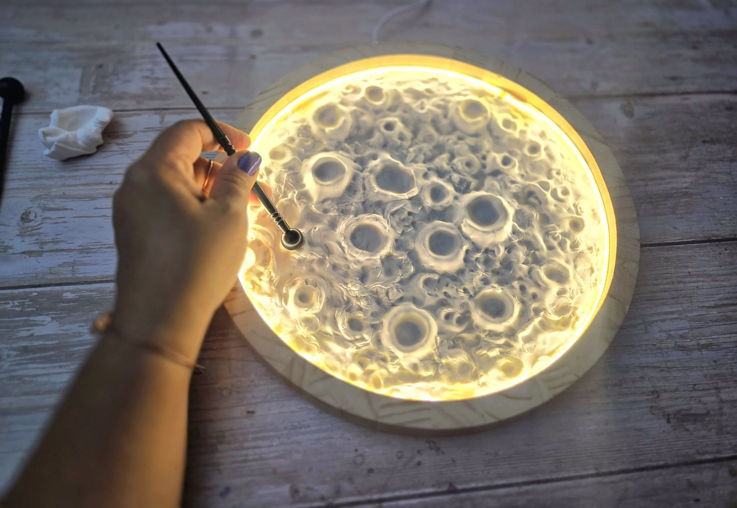 A glowy moon artwork is being created. A hand holds a stylus tool, creating craters within a round frame that is lit and looks like a moon.