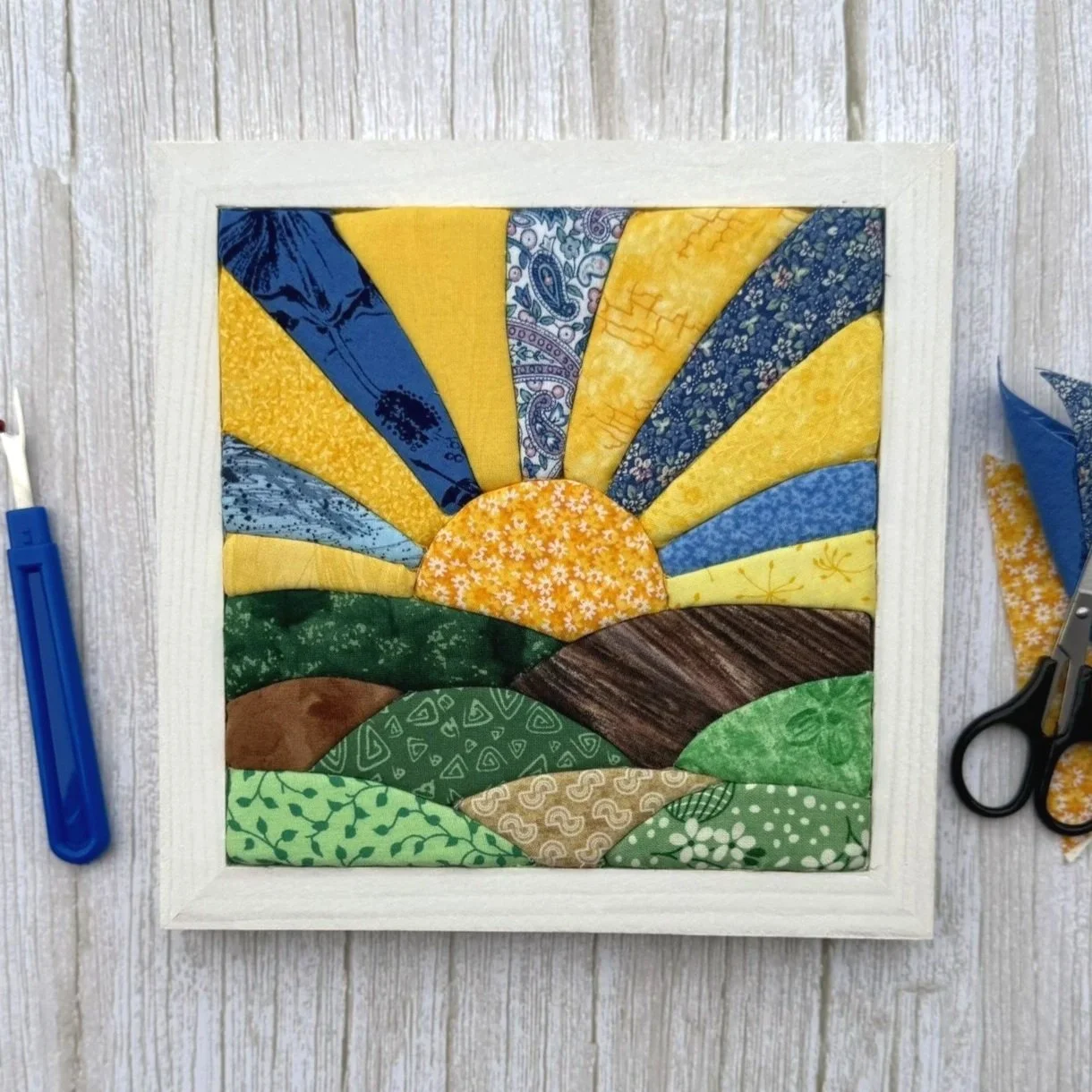 A framed kinusaiga piece, which resembles a quilt. It depicts a sunrise over rolling green hills with various patterned fabrics in yellow, blue, green, and brown.