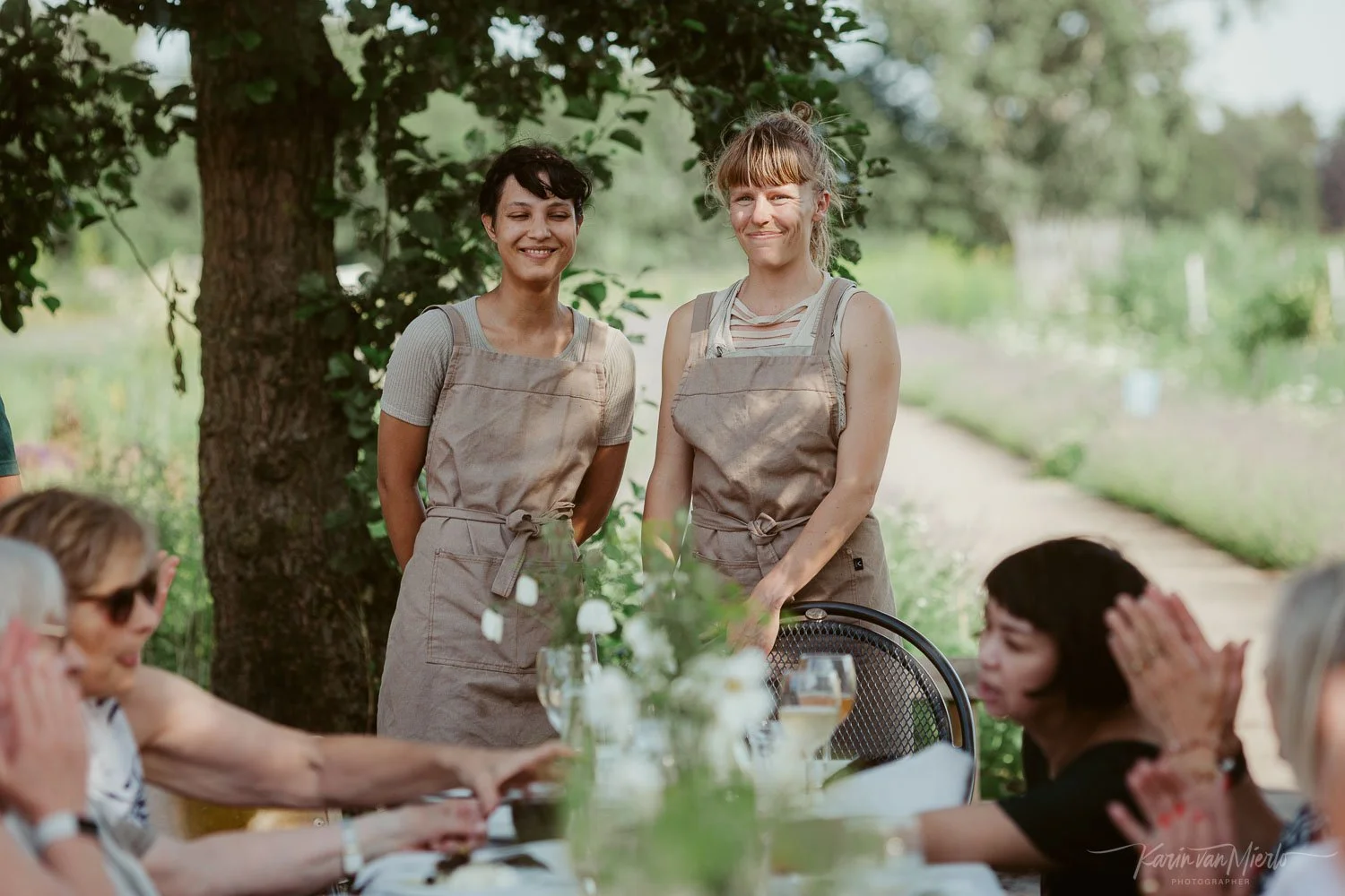 Twee vrouwen staan in een buitenrestaurant onder een grote boom, omringd door mensen die aan tafel zitten. De vrouwen dragen schorten en glimlachen naar de camera.