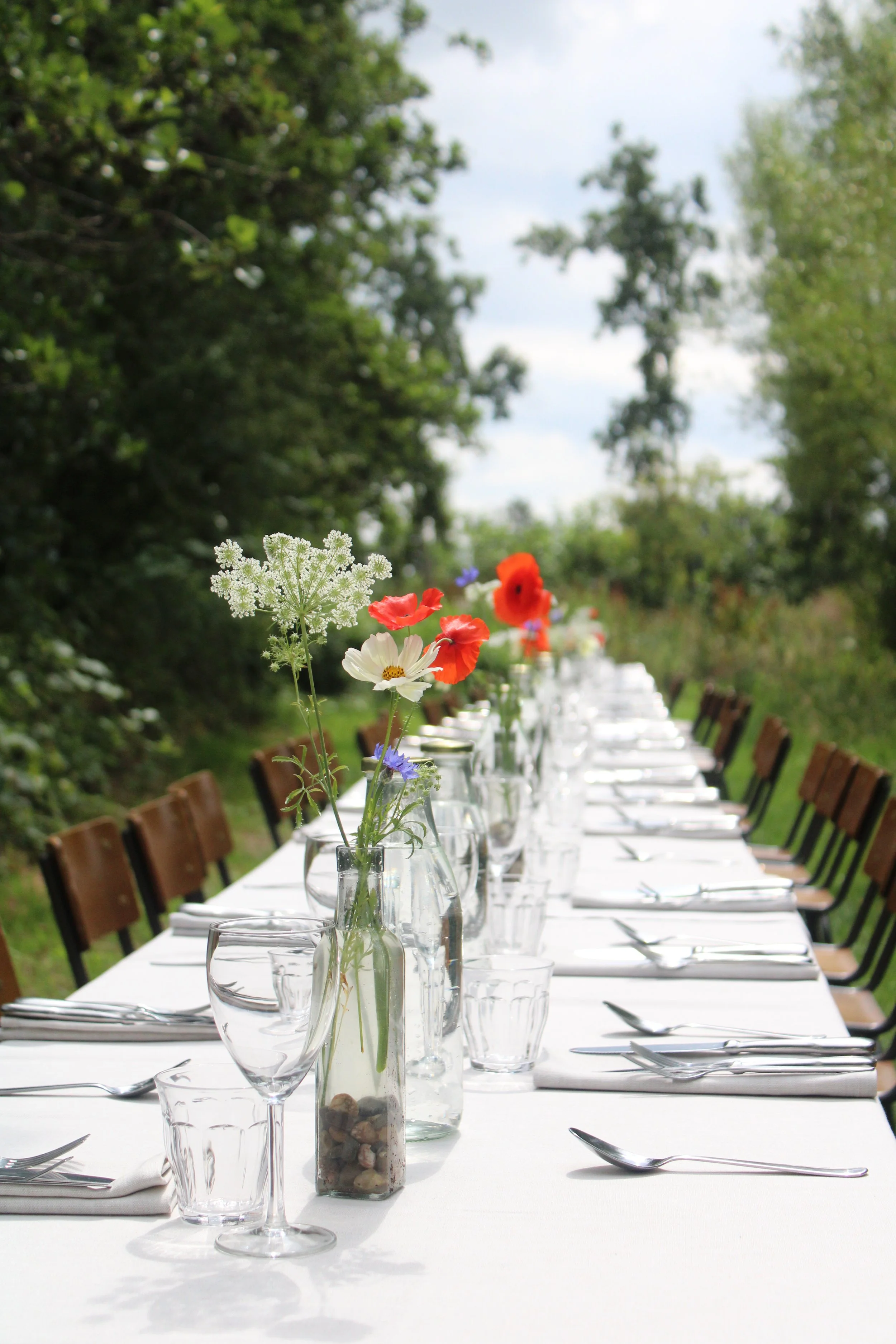 Lange tafel buiten met witte tafelkleed, stilleven met bloemen in vazen en set bestek, omgeven door groene bomen en een blauwe hemel.