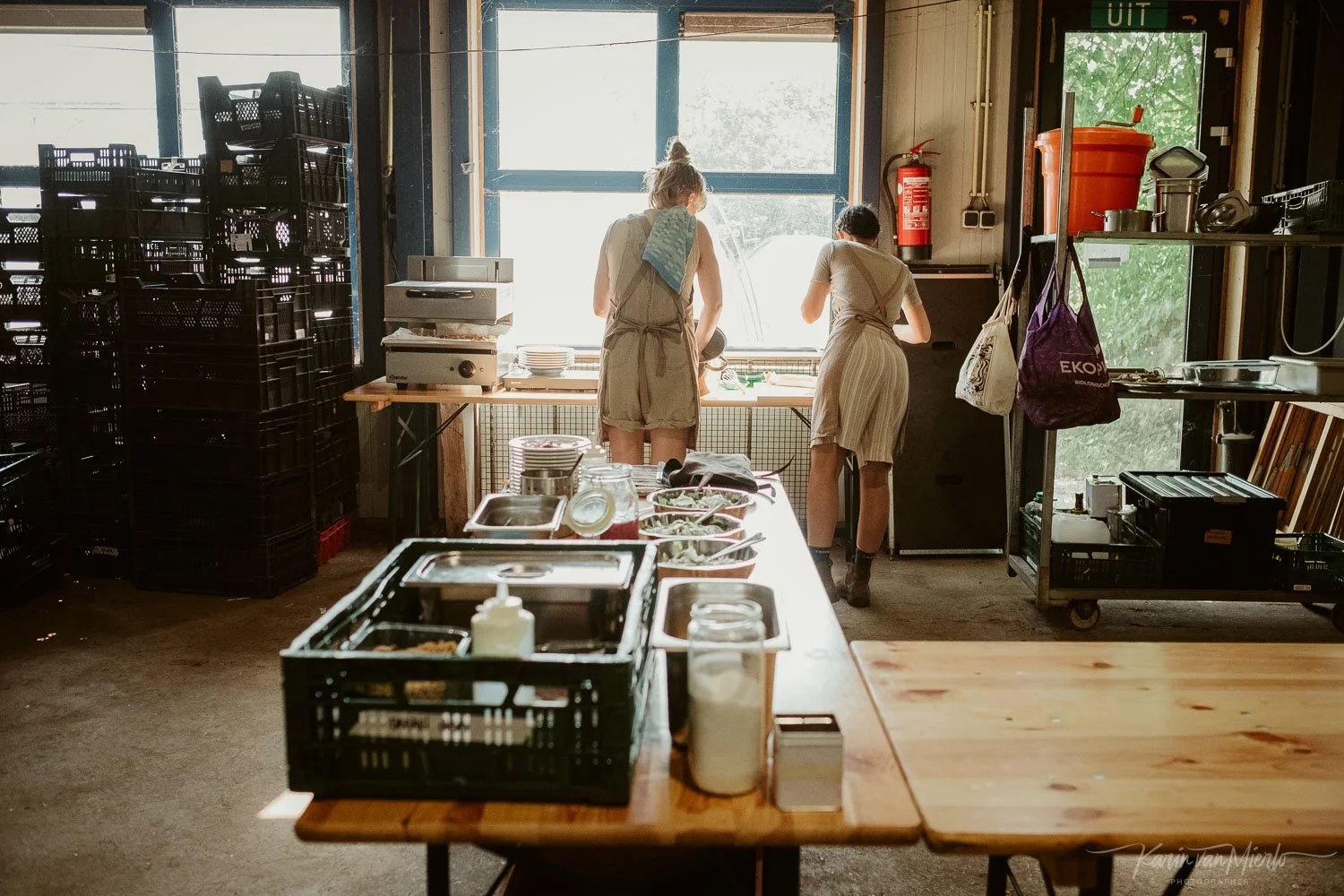 Drie vrouwen in een keuken of serverruimte, bezig met koken of bereiden van voedsel, met ramen en diverse keukenbenodigdheden.
