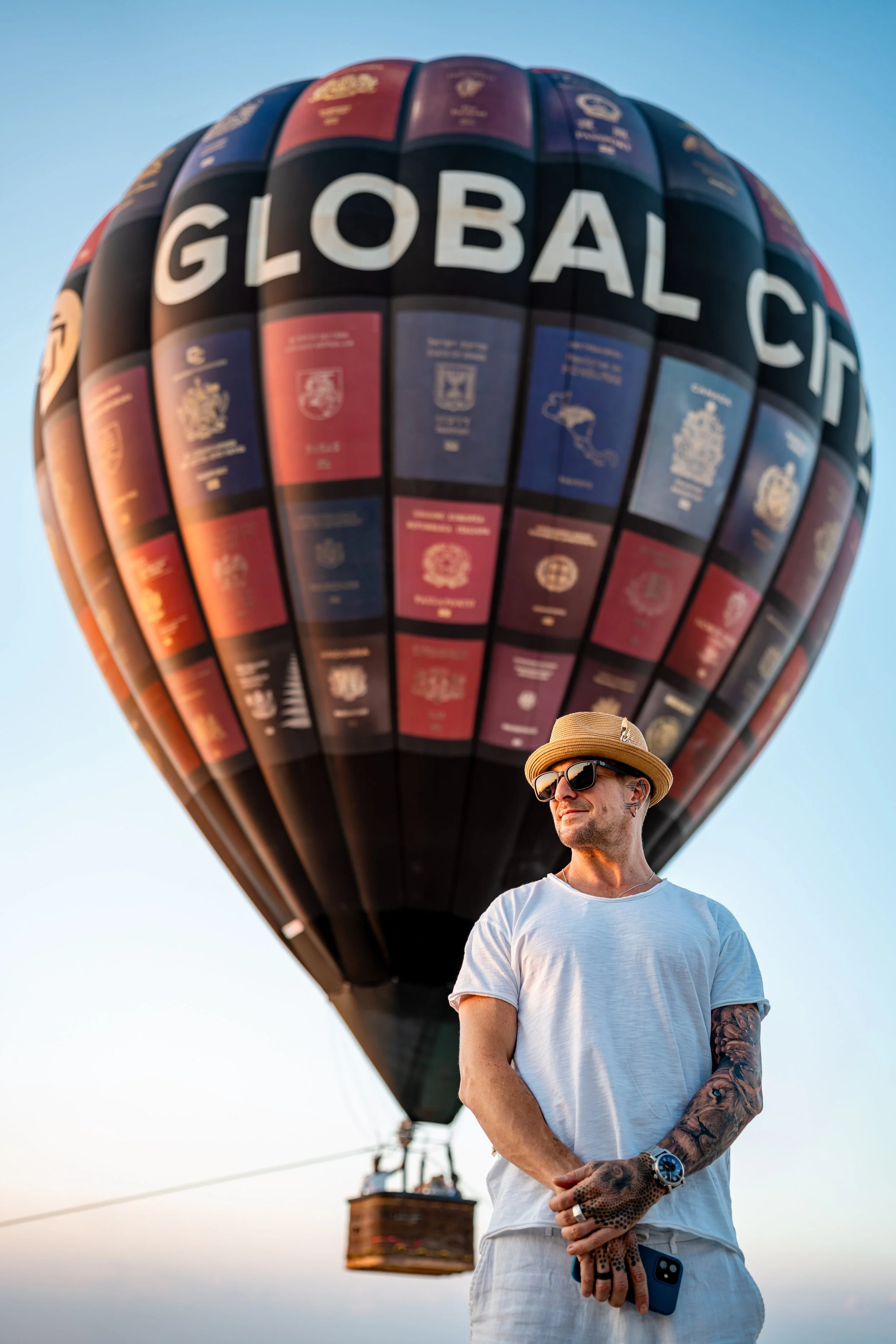 Man wearing sunglasses, a hat, and a white t-shirt stands in front of a hot air balloon with the words "Global City" on it, during sunset.