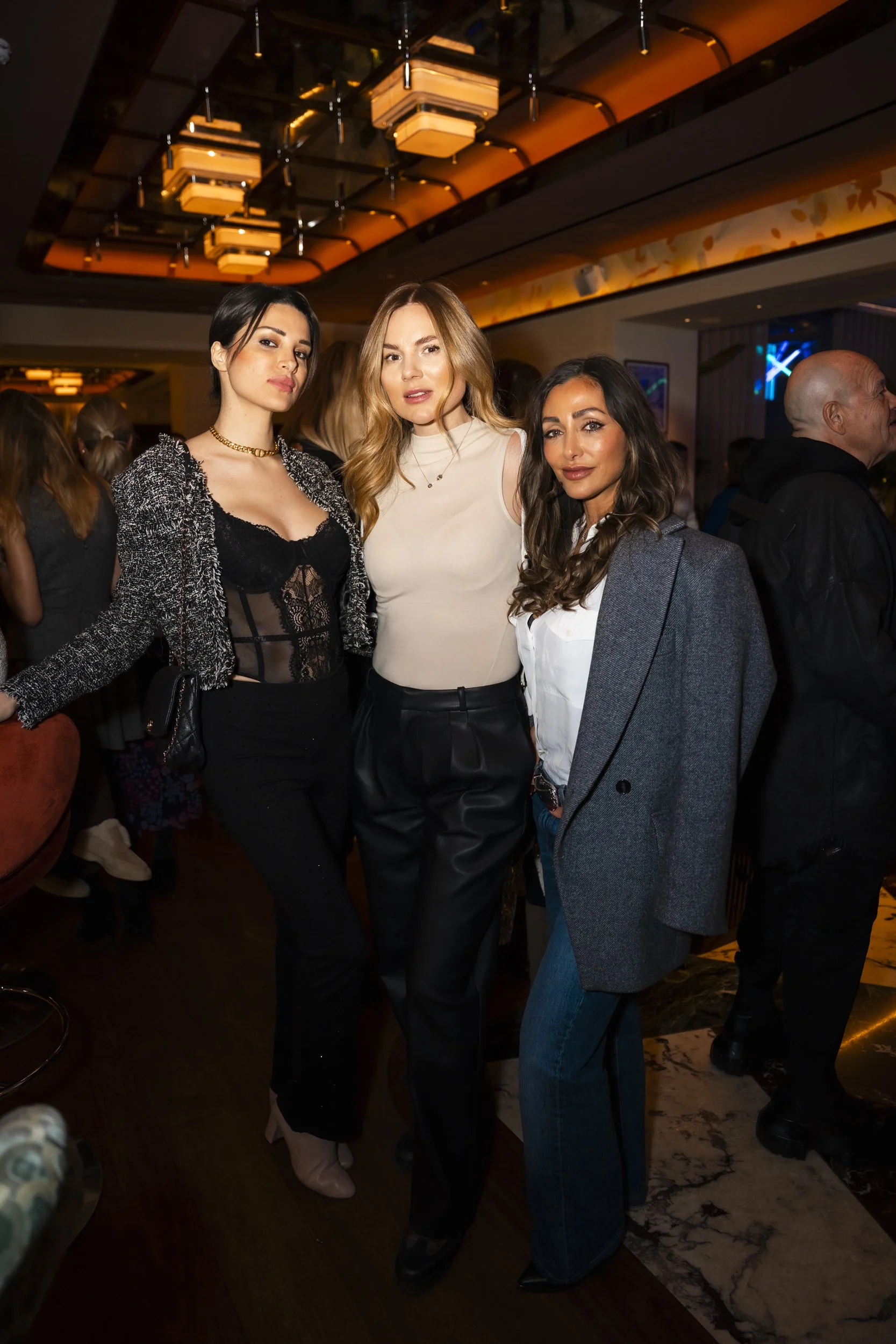 Three women posing together at a social event, with a warmly lit interior and many other people in the background.